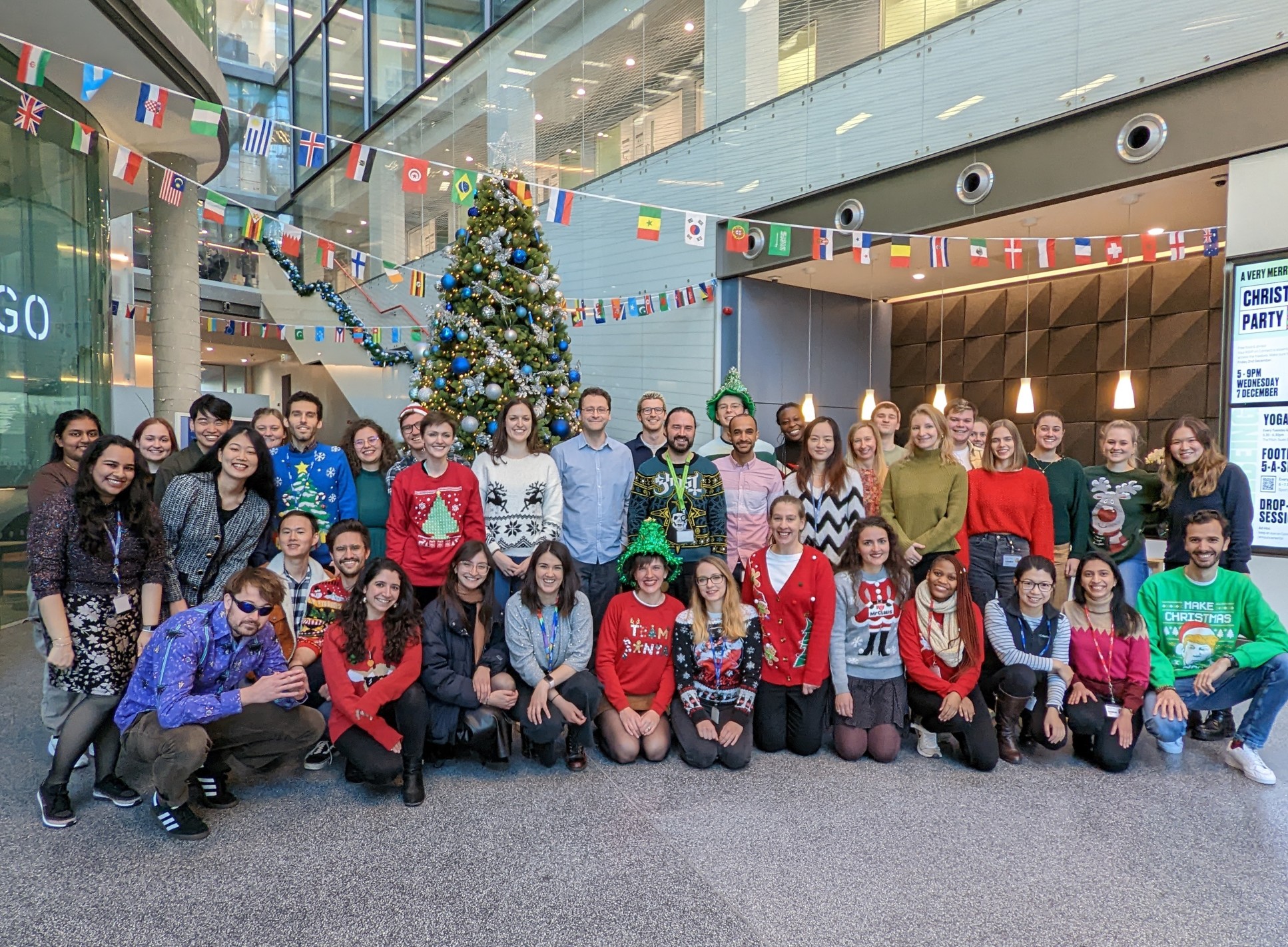 A large group of people wearing Christmas jumpers in front of a Christmas tree and flags of the world