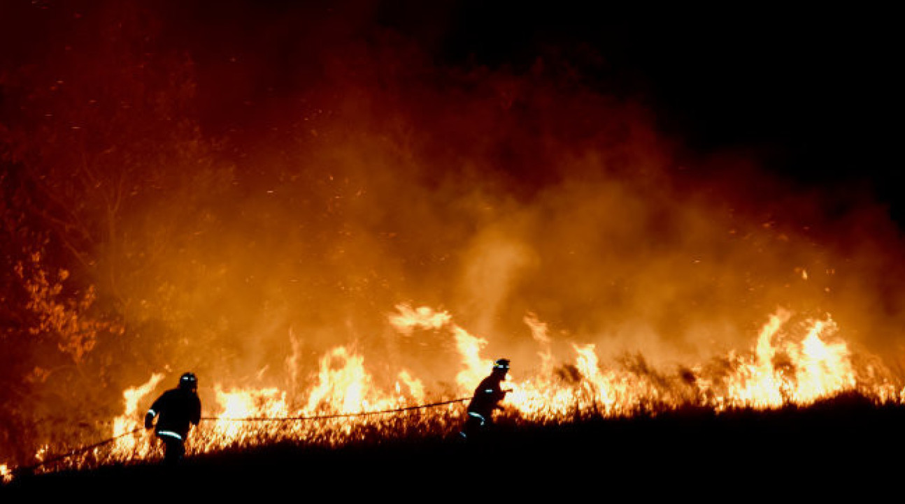 Two fire fighters pull a hose through grasslands as a fire blazes twice their height