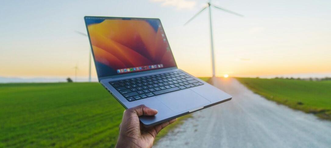 A hand holding up an Apple Macbook and wind turbines in the background