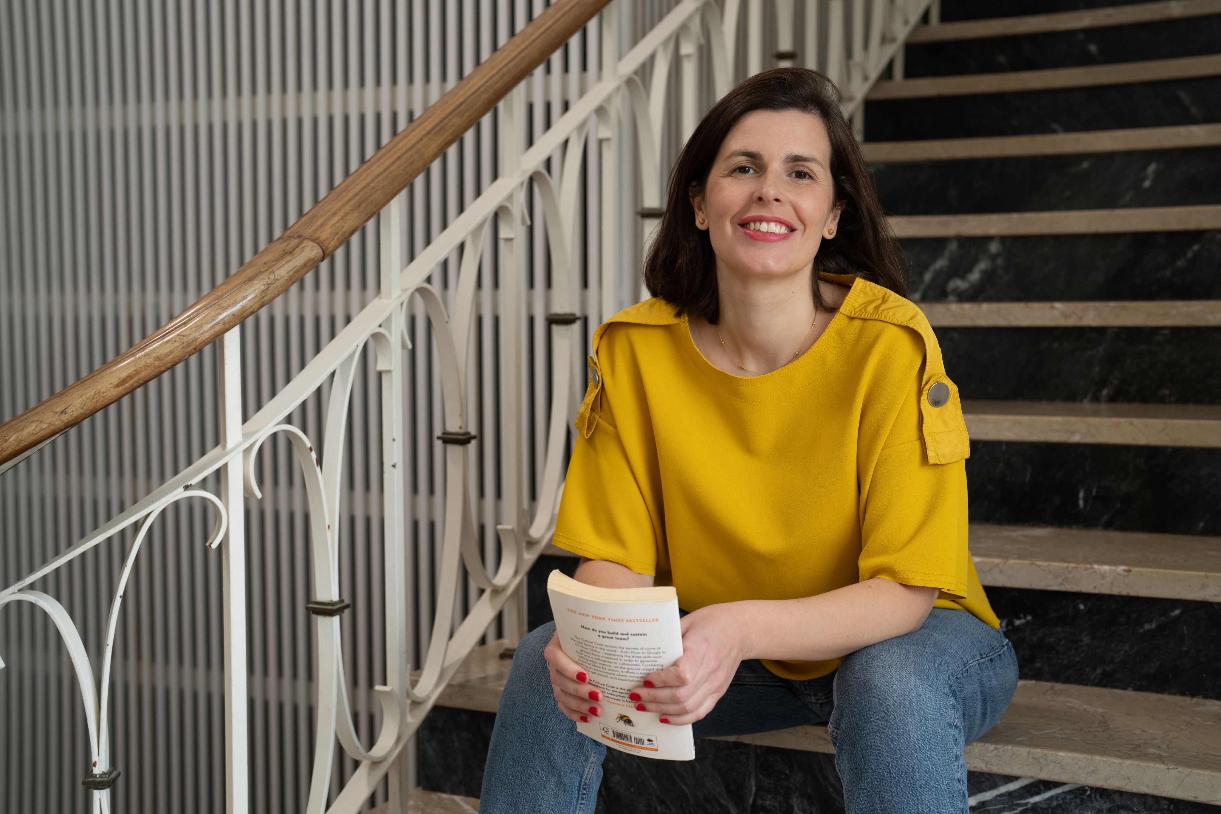 Joana Moscoso wearing a yellow top, holding a book sat on a staircase