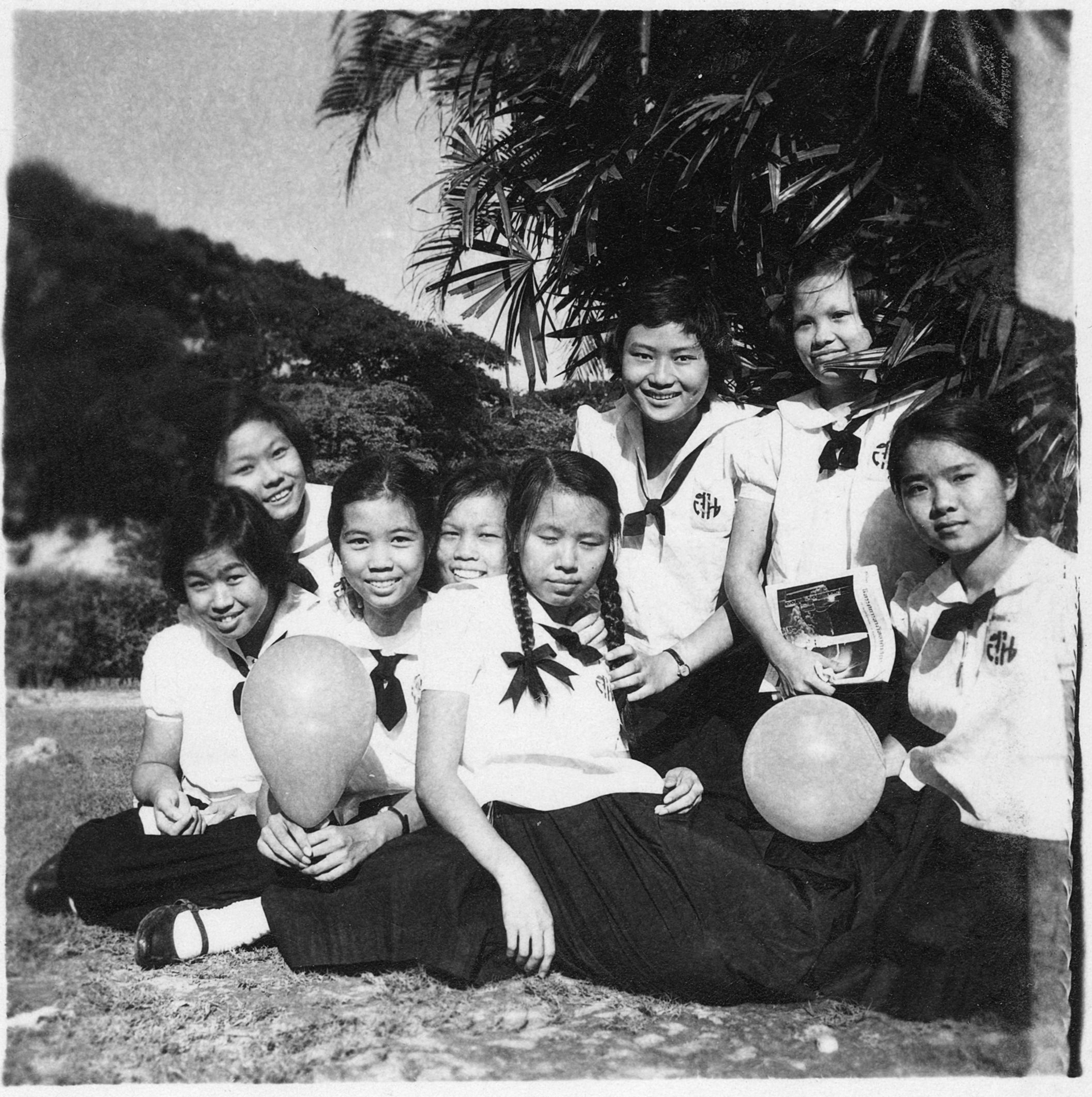 A black and white photo of a group of 8 Thai girls in school uniform sat in a group under a tree