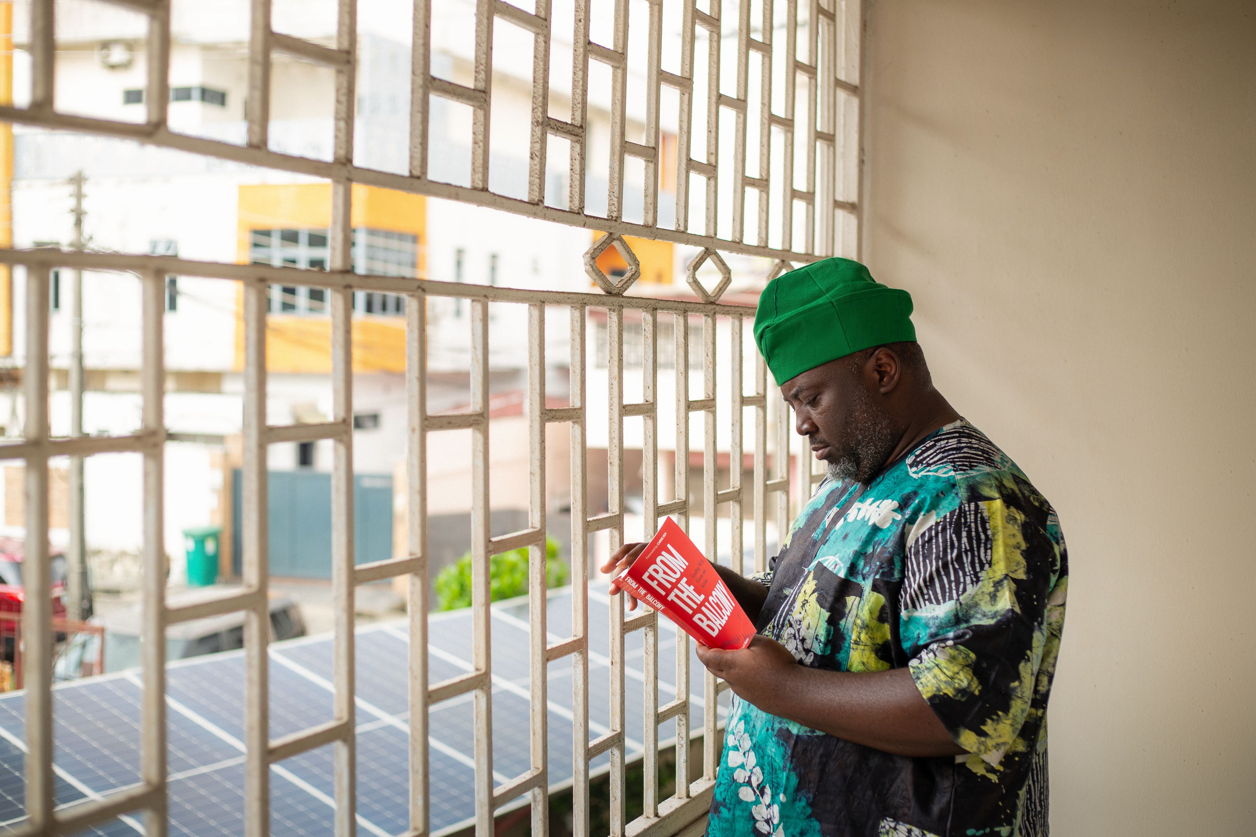 Kolawole Osinowo wearing a green fila cap and a matching patterned Ankara short-sleeved top and trousers standing indoors by a window reading a book titled 'From the balcony'