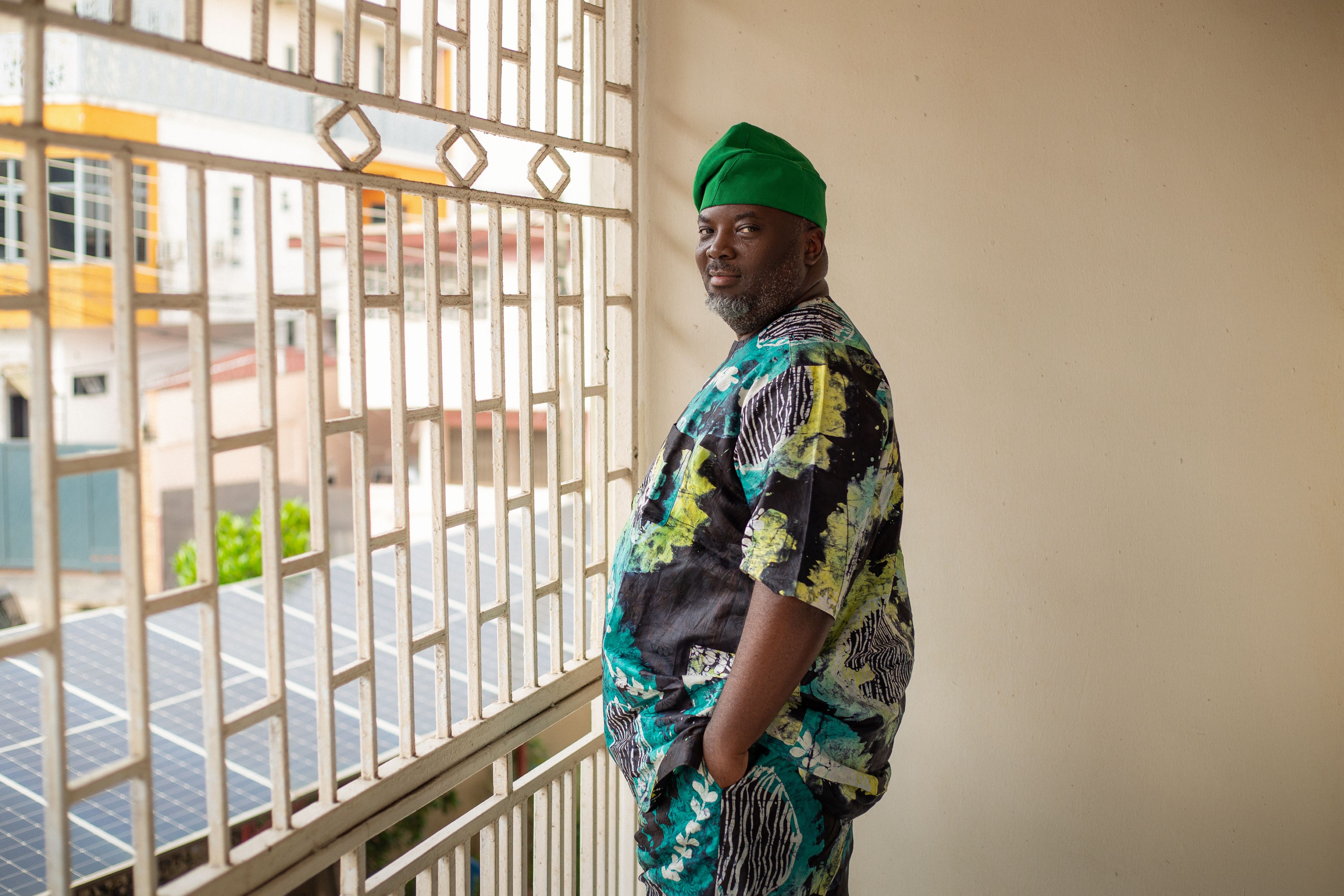 Kolawole Osinowo wearing a green fila cap and a matching patterned Ankara short-sleeved top and trousers standing indoors by a window