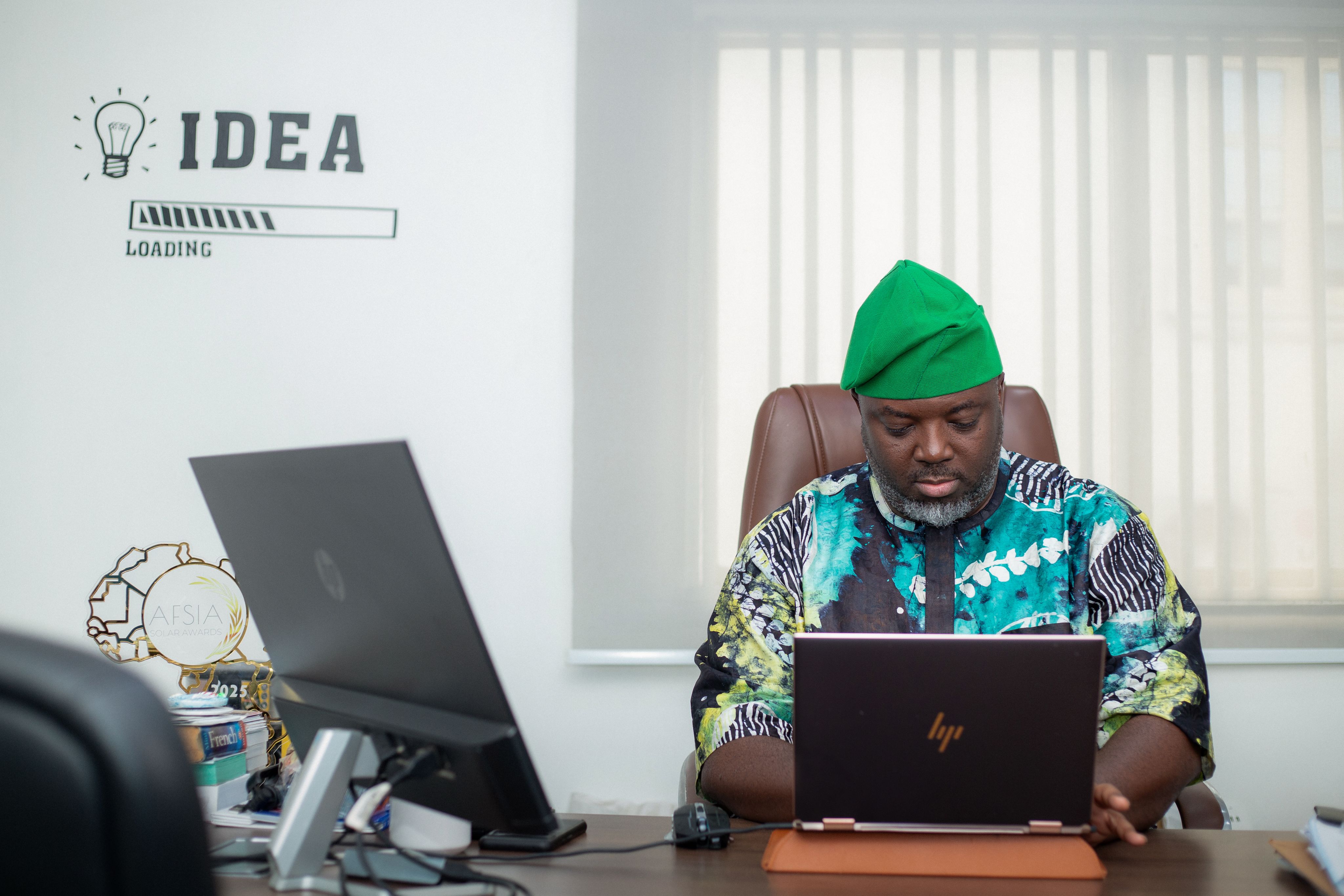 Kolawole Osinowo sat at his desk wearing a green fila cap and a matching patterned Ankara short-sleeved top and trousers
