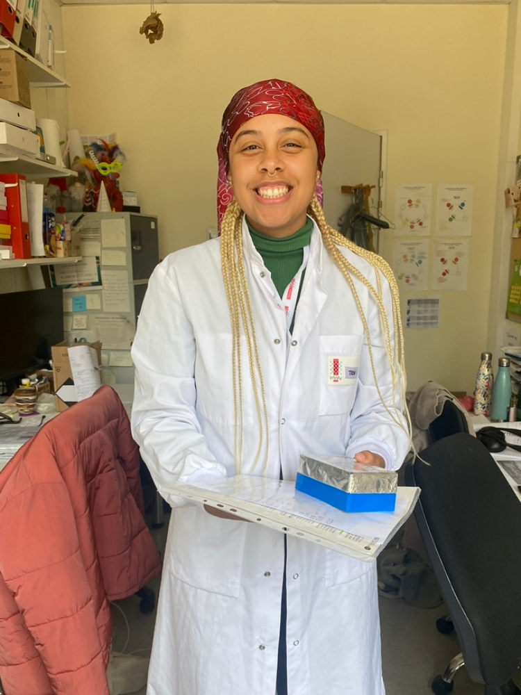 Tasha smiles to the camera whilst wearing a white lab coat and holding a folder and her pencilcase