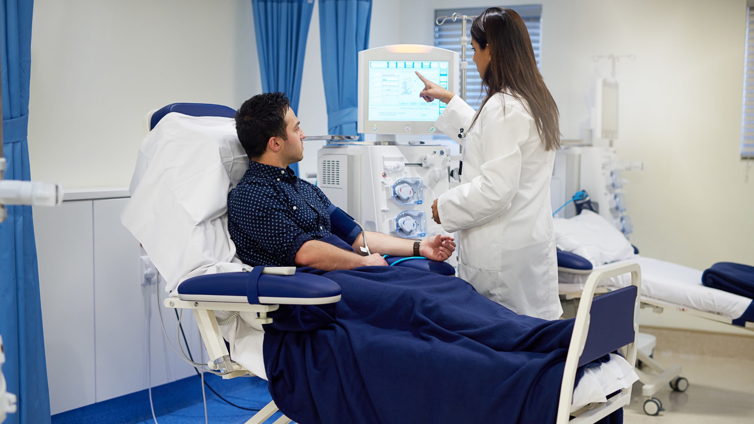 A male patient in a hospital bed is shown medical data on a screen by a female doctor