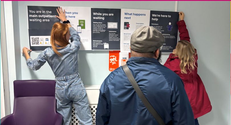 Two women pin information on a wall, a man watches