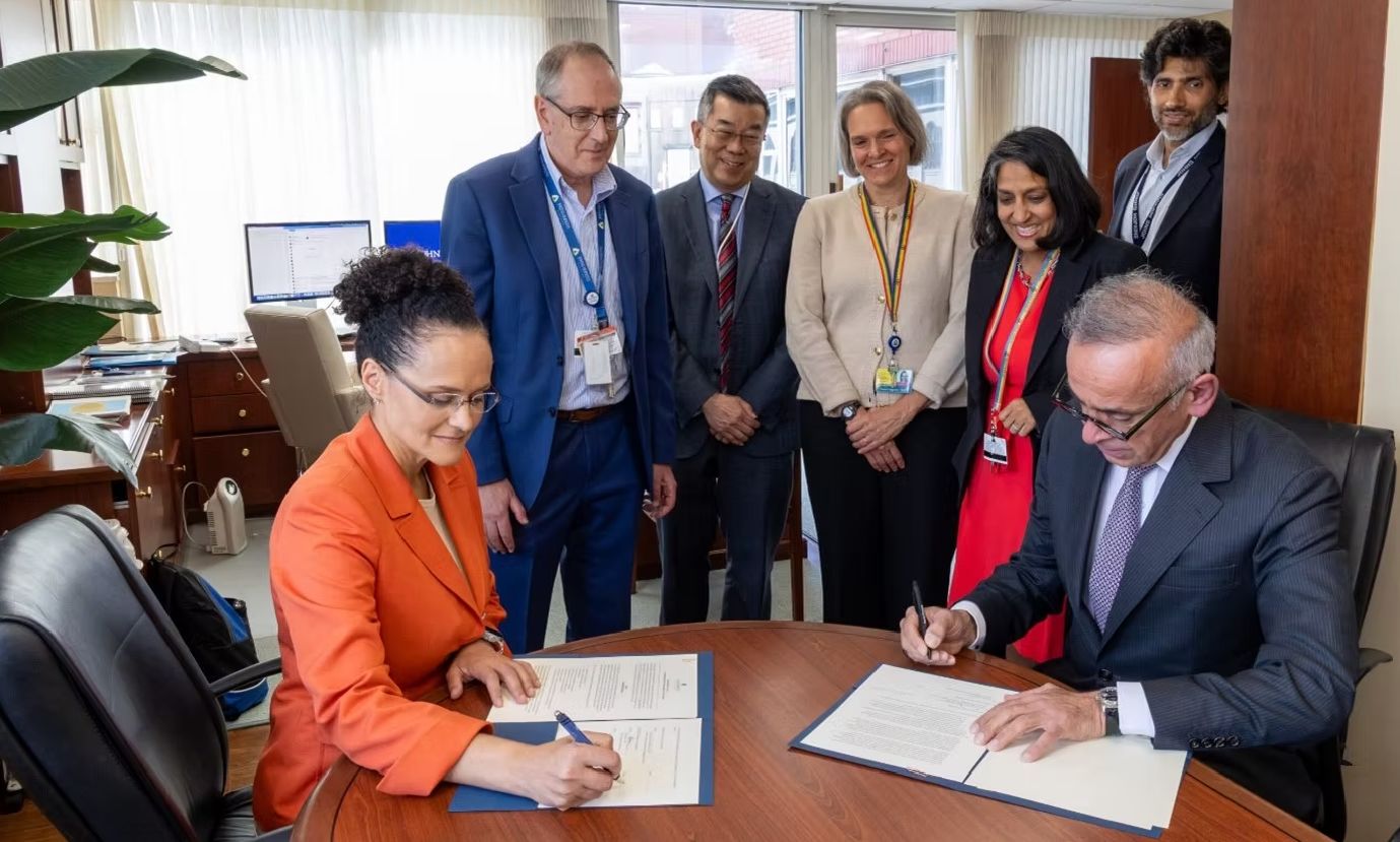 A man and a woman seating, signing documents. Five people standing watching
