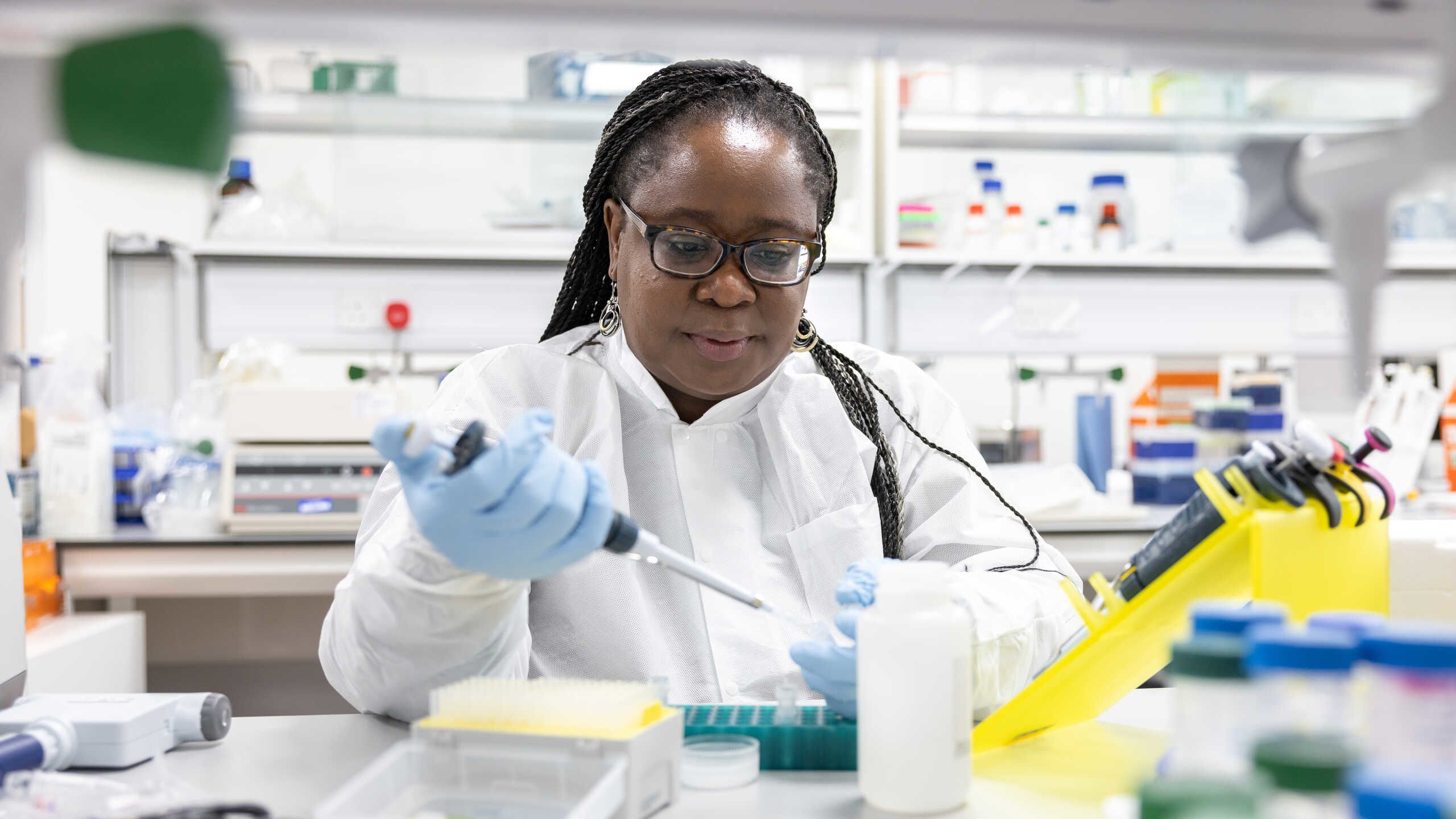Dr Catherine Kibirige in the Department of Infectious Disease at the Chelsea and Westminster Hospital. She wears a white lab coat and holds a pipette in one hand.