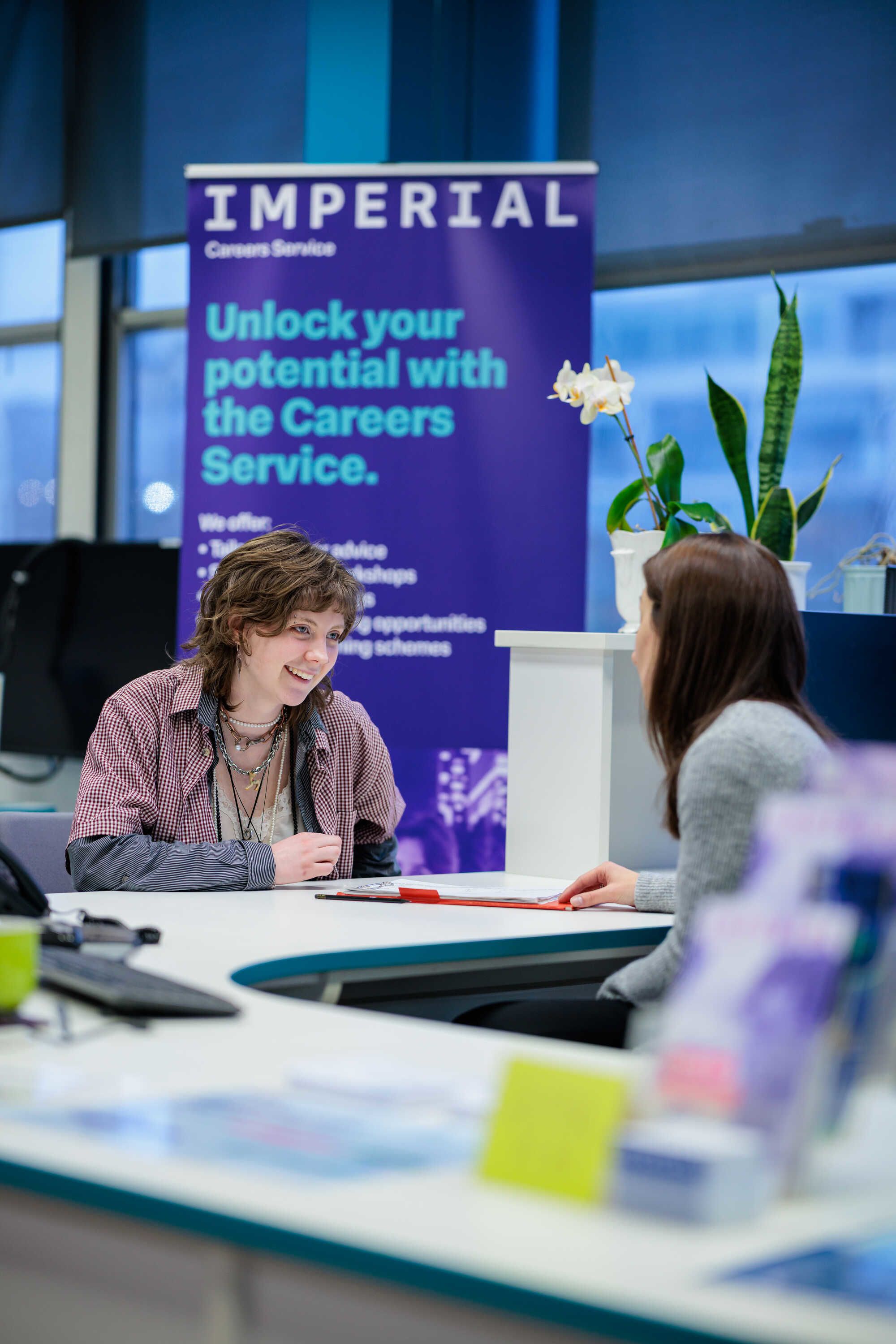 Two people sat at a desk talking with a pop-up banner behind them with an Imperial logo and "Unlock your potential with the Careers Service"