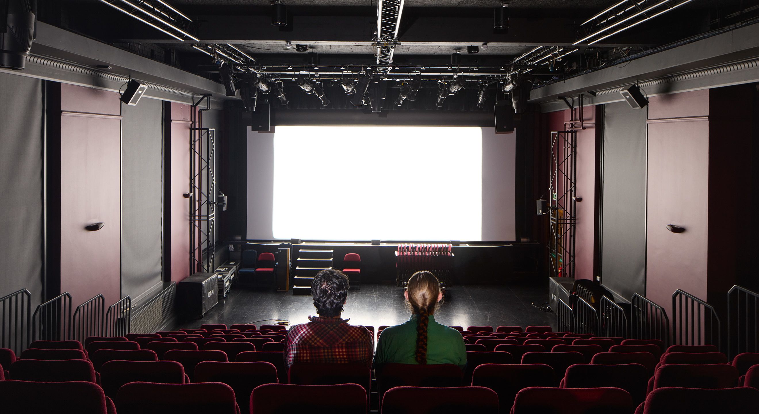 Two people sit in a dark room and look at the blank projector screen belonging to the Imperial Cinema Club