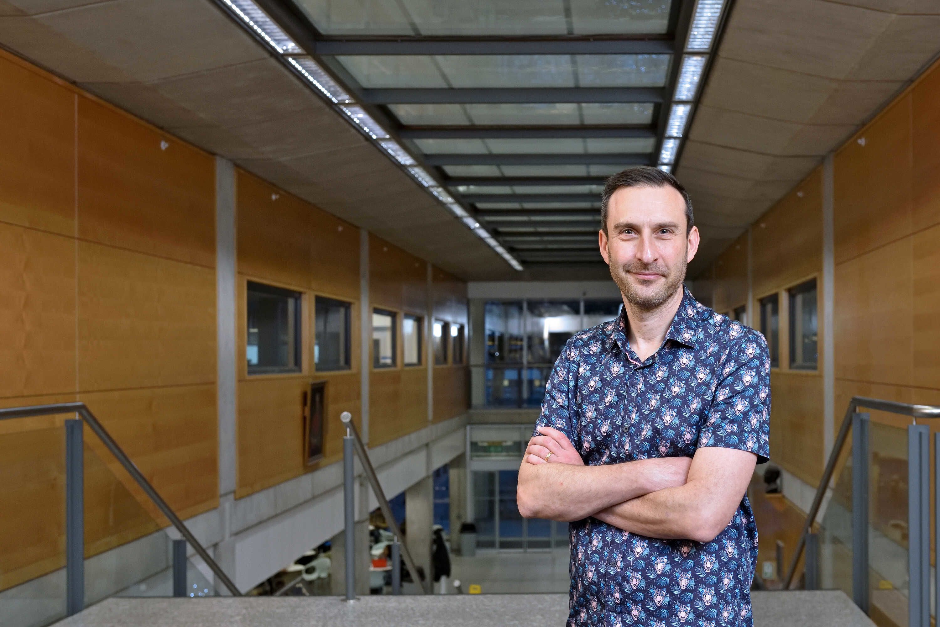 Simon Foster with his arms crossed staring sternly into the camera standing at the top of a stair case in the Sir Alexander Flemming building.