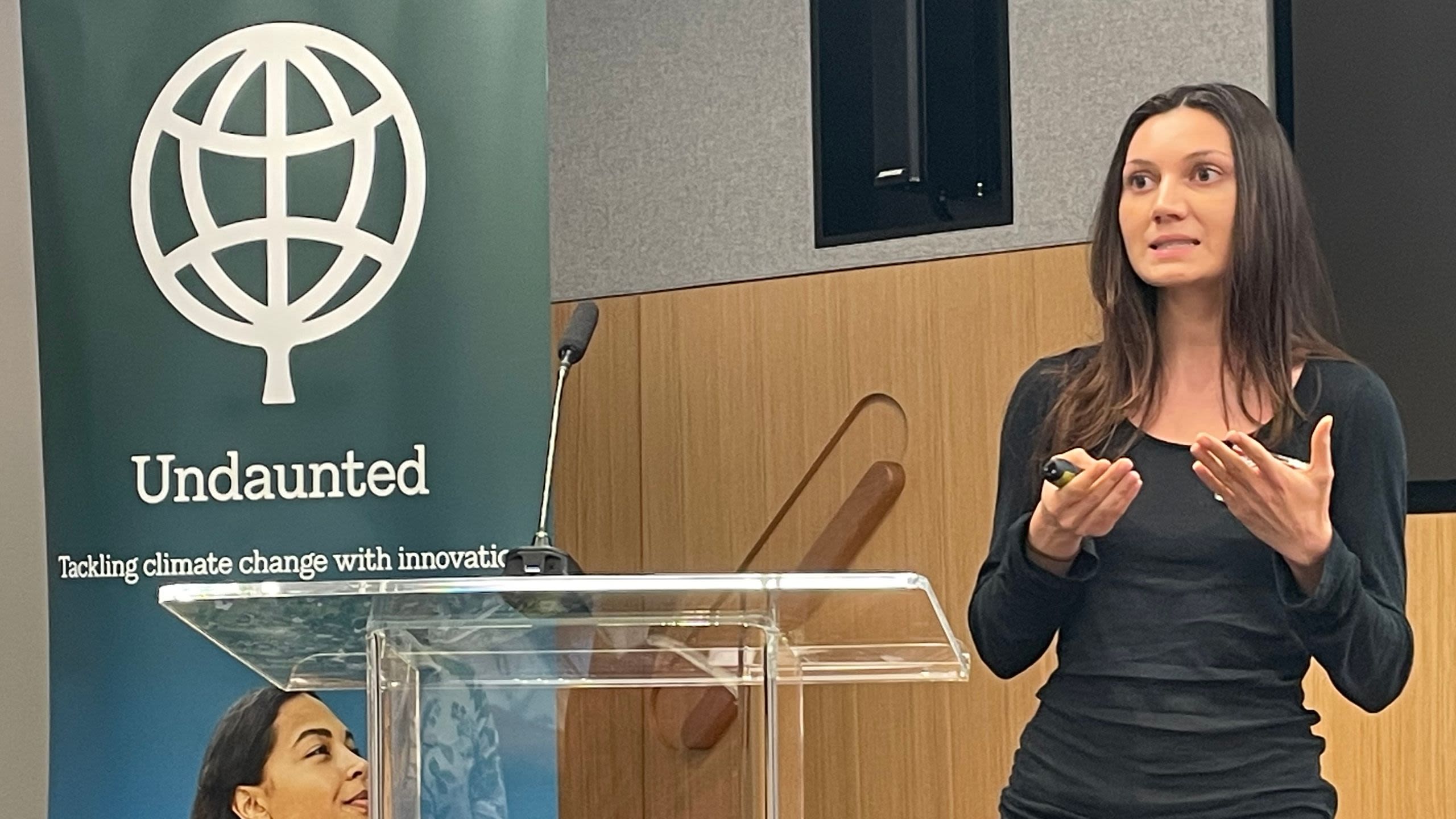 Mara speaking at an event by a lectern and a branded Undaunted banner
