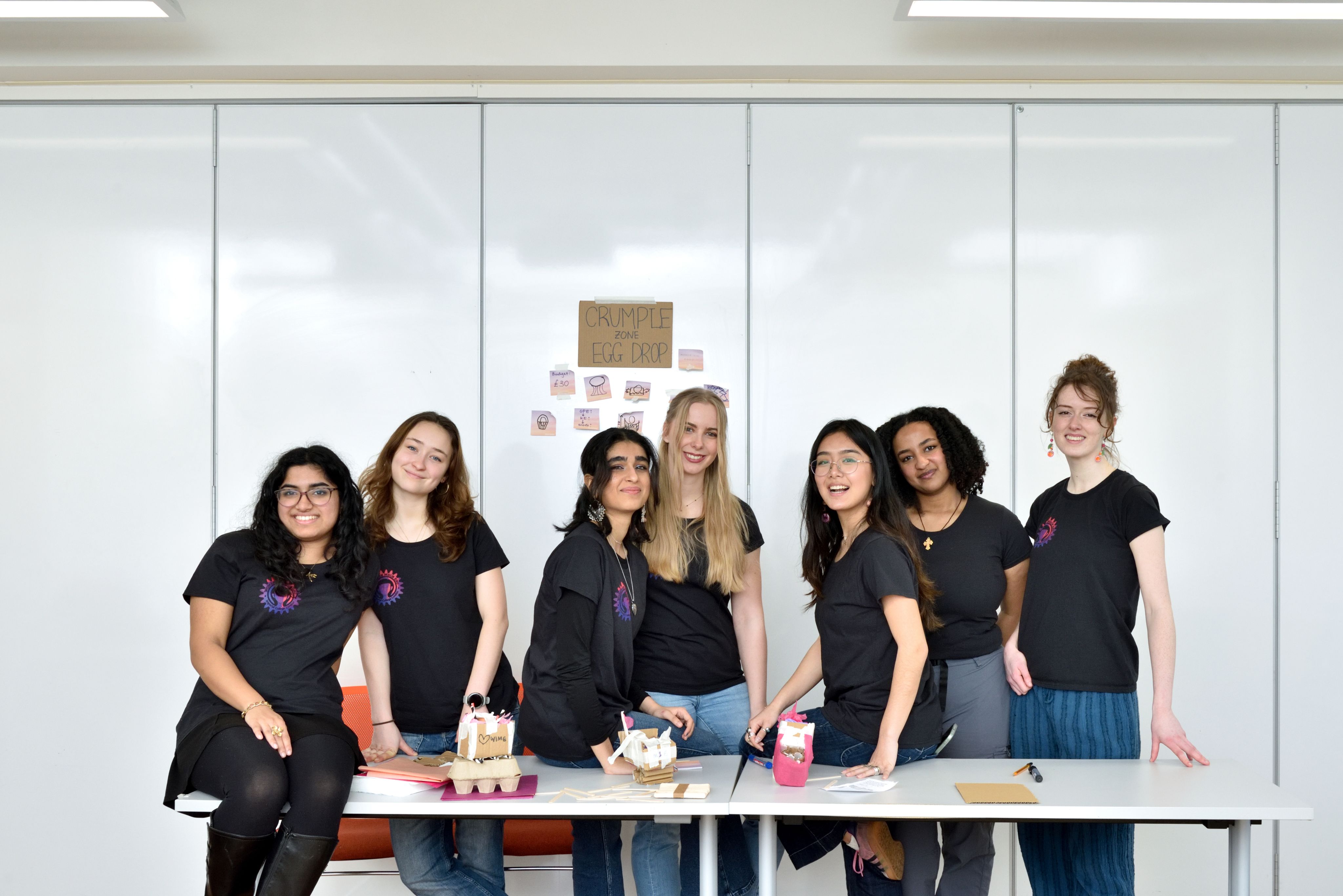 Seven members of the society together standing behind a workshop table and smiling at the camera, there is an crumple-zone egg drop sign behind them