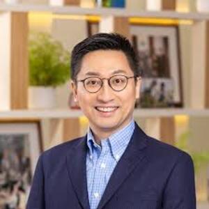 man standing in front of book shelf. Wearing glasses and a jacket and checked shirt.