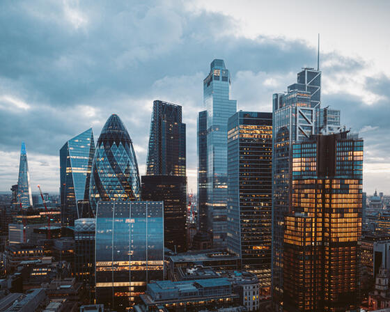 A collection of skyscrapers in the London skyline shows buildings with some lights on, in front of a grey, cloudy sky