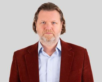 The photograph shows a man with long hair standing and looking straight into the camera. The image is cut off at his waist. he is wearing a dark red blazer and light blue shirt. 