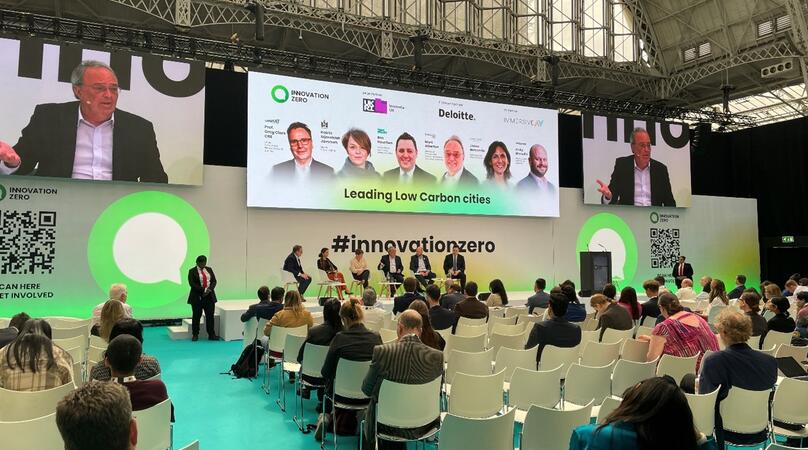 A panel of speakers sit on stage, discussing in front of a large audience, with event graphics behind them