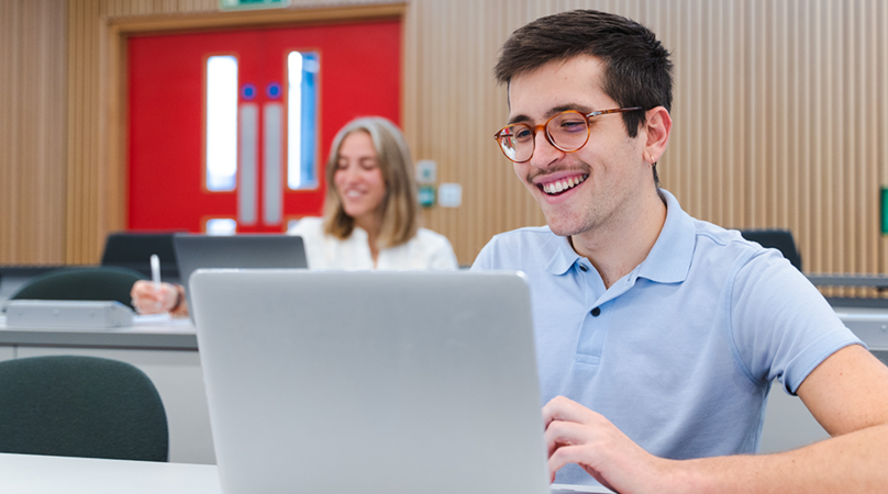 A young male student with glasses on a laptop smiling in a lecture room