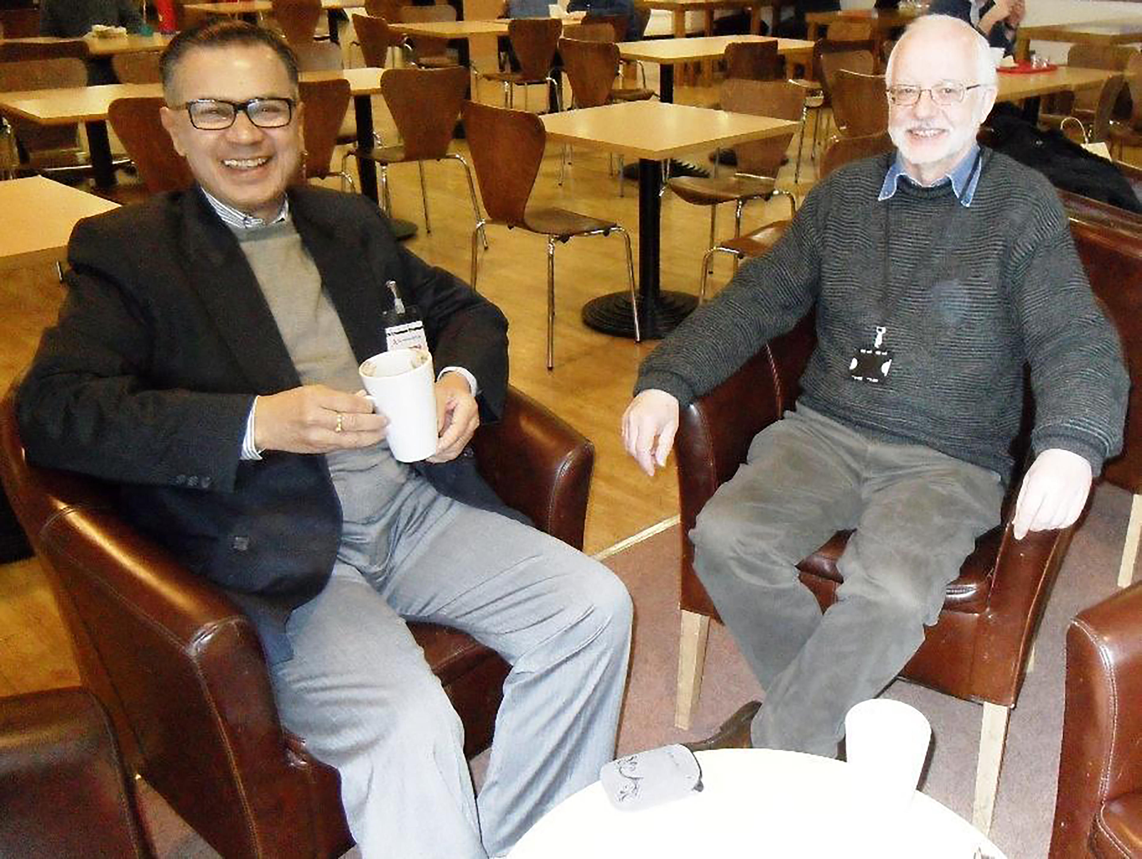 Dr Hirak Sen and Peter sitting back in armchairs at a cafe. Hirak is holding a mug. They are both smiling at the camera