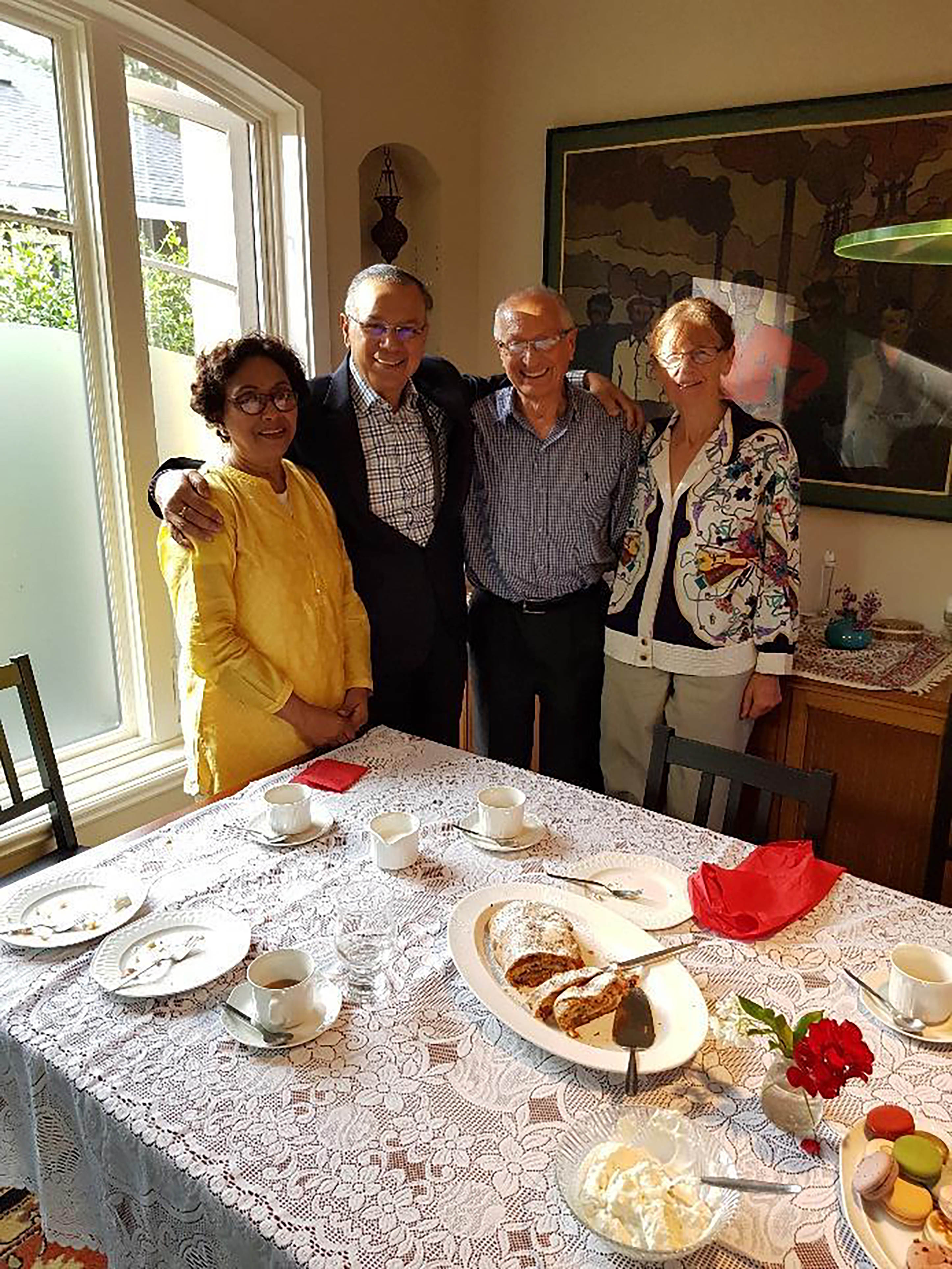 The group are standing around a table of food and empty dessert plates, there are cakes/sweets/cream in the middle. The room is sunshine yellow and there is a large window behind them.