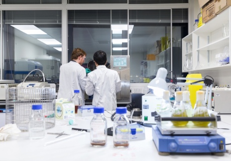 Two men in lab coats working on a lab bench behind flasks of green liquid