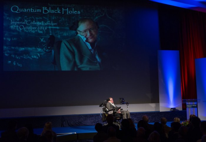 Stephen Hawking on stage in front of a large title slide for the lecture