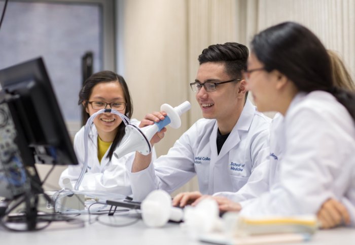 three students in white coats, one holding up a medical instrument