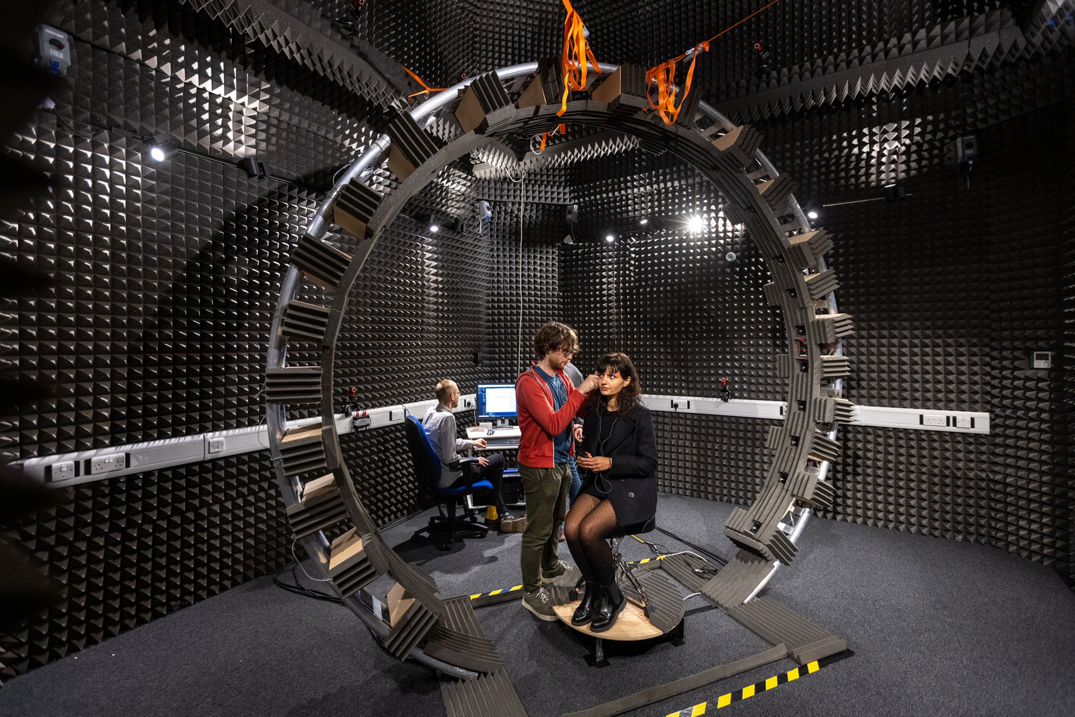 A volunteer sits in a rig as a researcher attaches audio devices in an audio experience suite