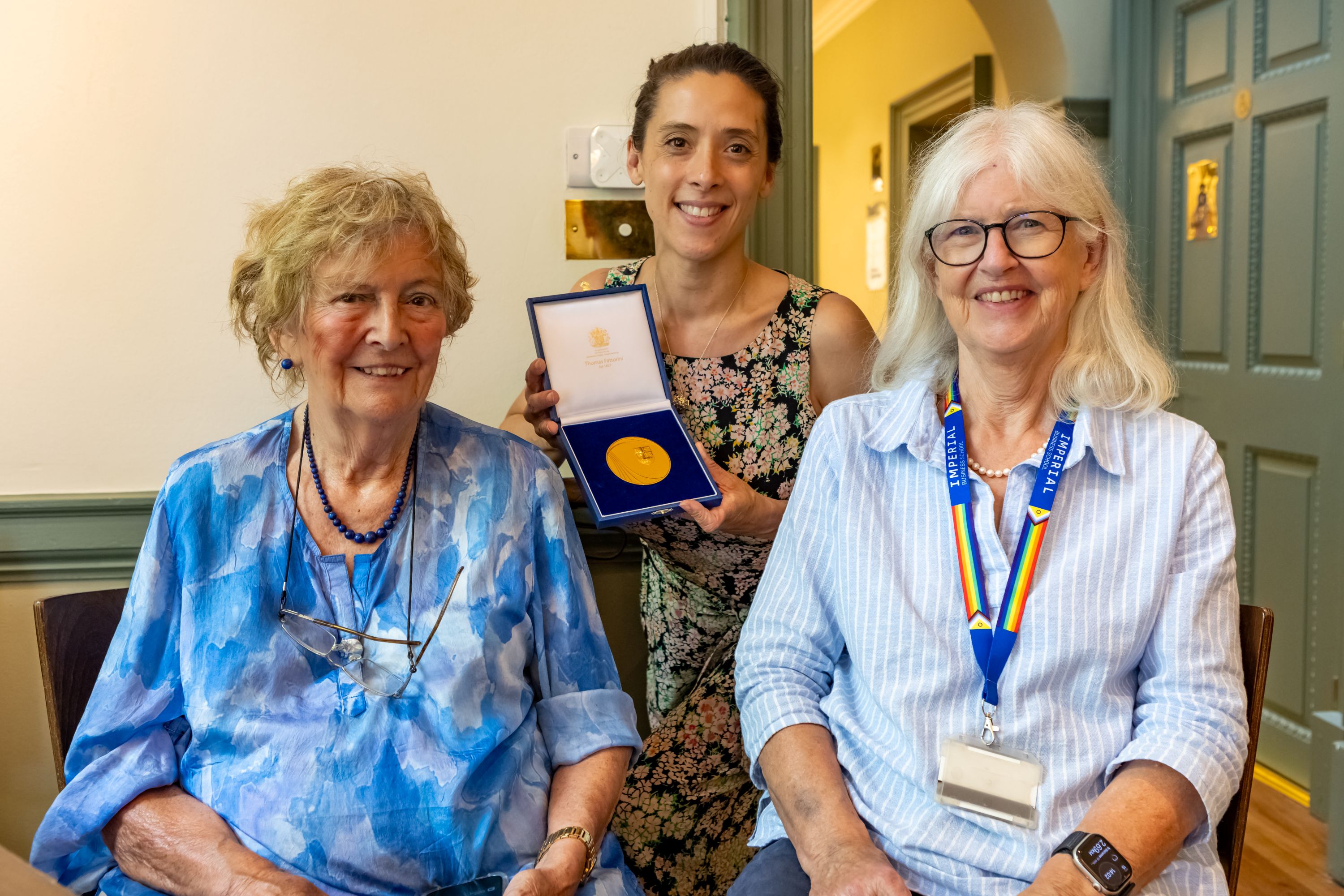 Three people pose indoors, with the person standing in the centre holding an open presentation case displaying a gold medal.