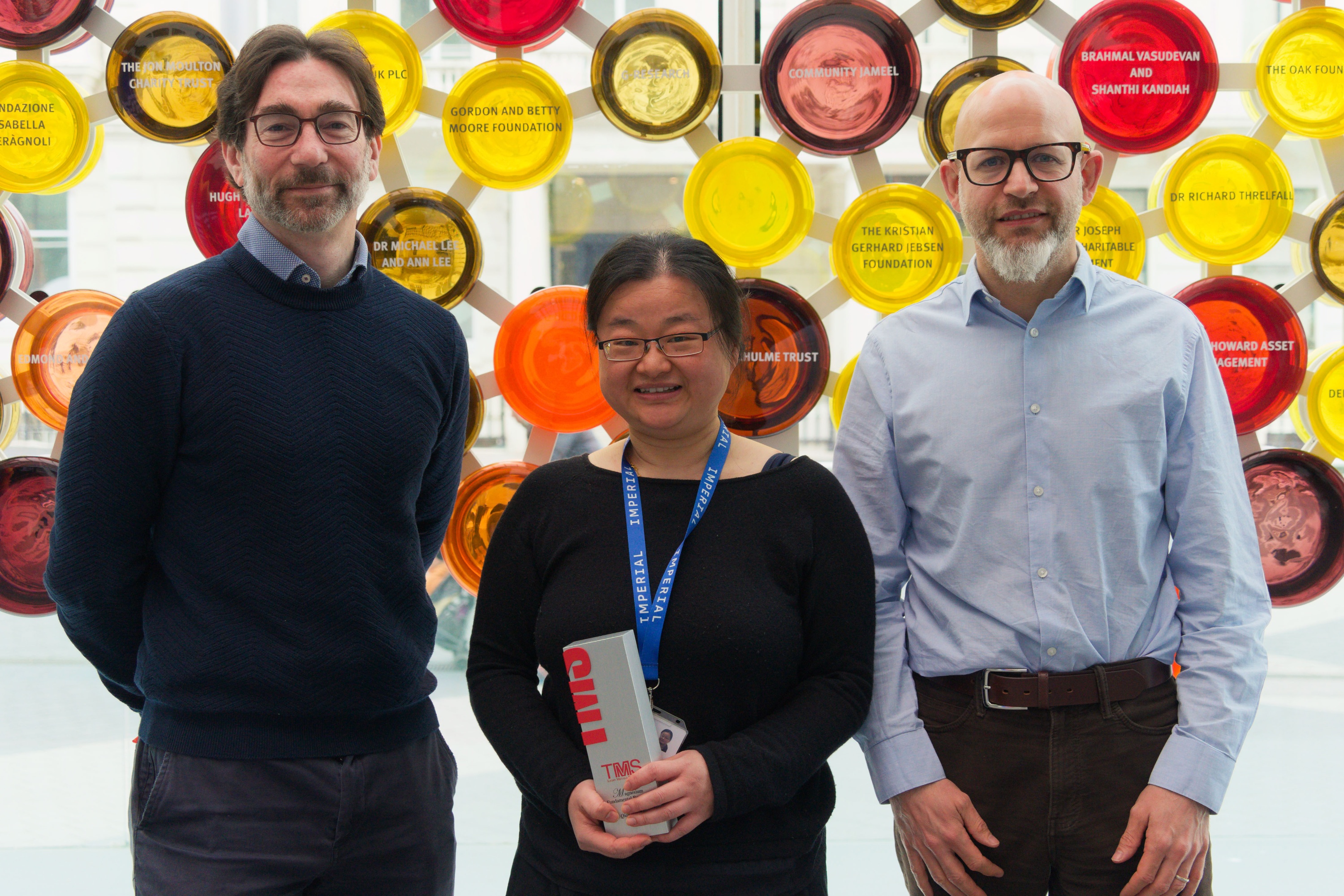 Milo Shaffer, Qianqian Li and Christopher Gourlay standing in front of an abstract background. Qianqian (middle) is holding the TMS award won by the team for their work on lightweight magnesium materials.