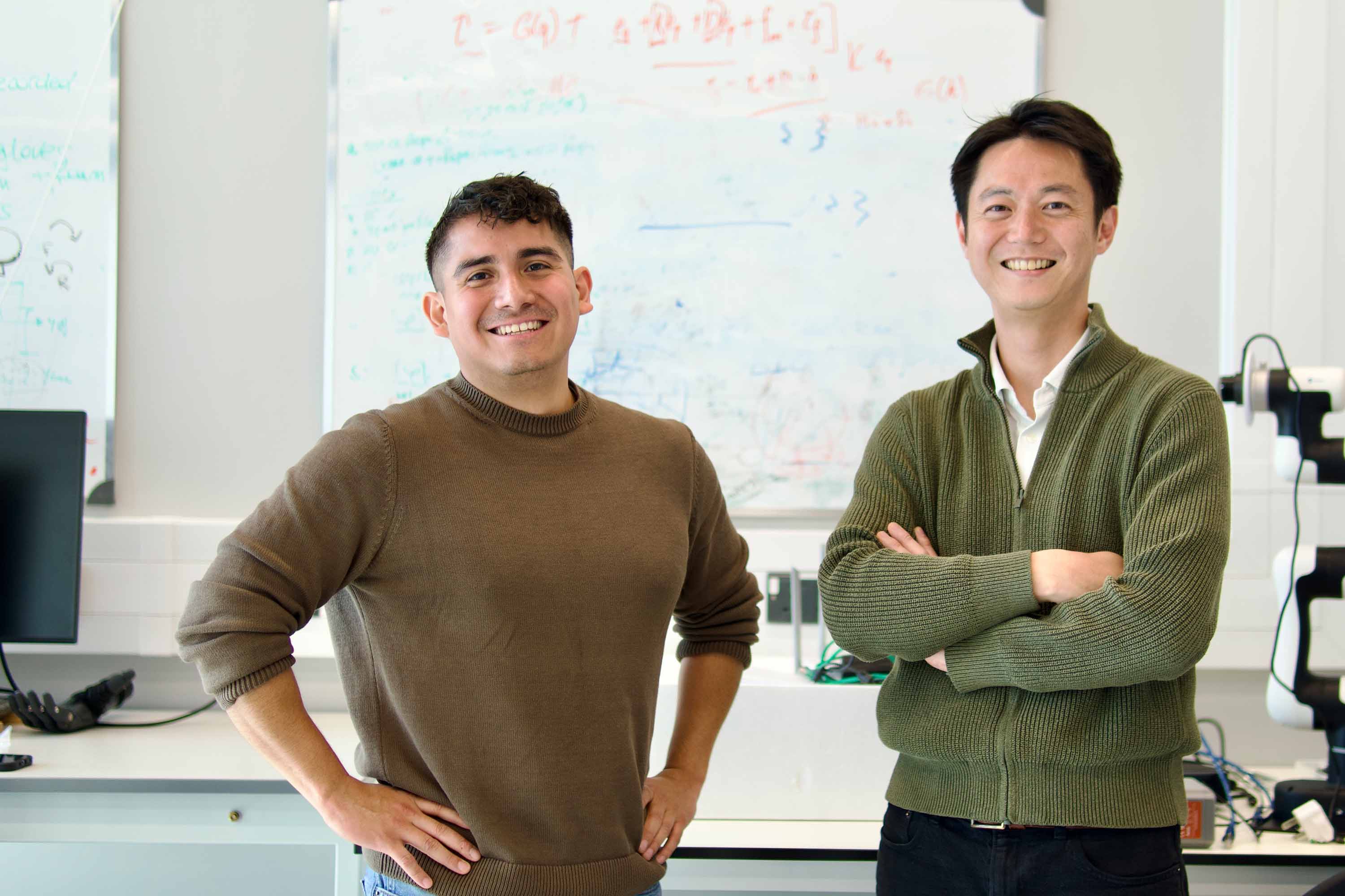 The founders of Neubond, Dr Patrick Sagastegui Alva (left) and Mr Jumpei Kashiwakura (right) stand in a laboratory. A whiteboard behind them is covered in equations, and to the right is a robotic arm.