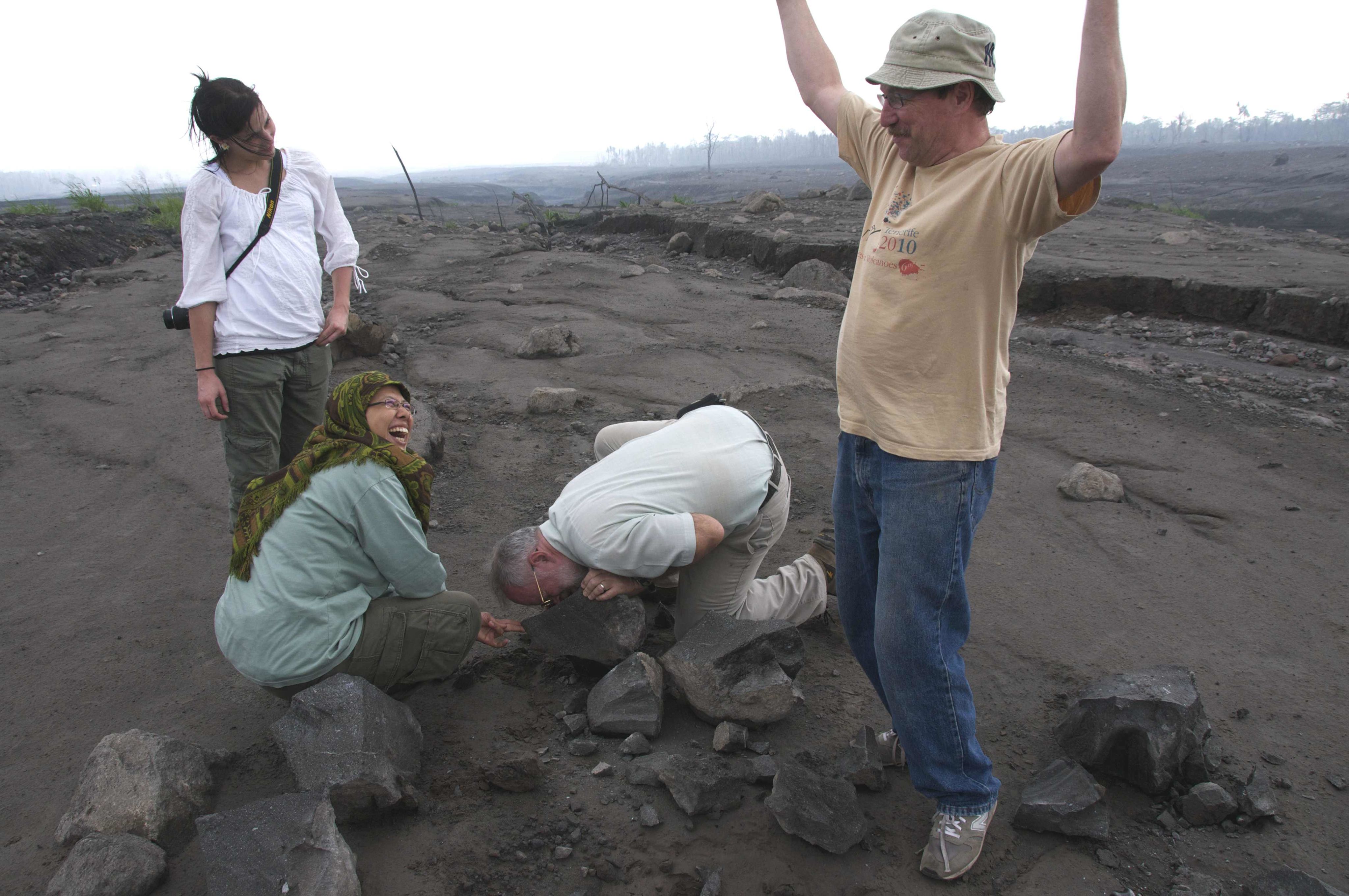 STephen stands on a rocky field with his arms in the air surrounded by laughing colleagues