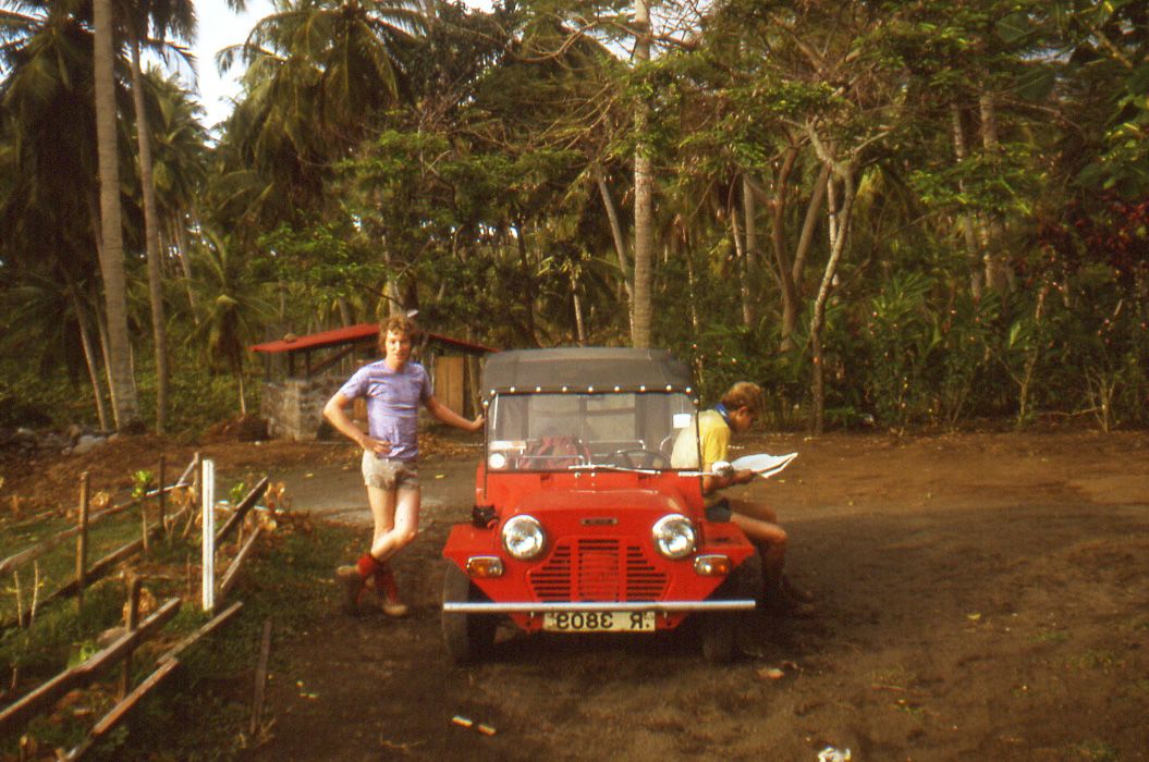 Stephen wears shorts and a tshirt, leaning against a red car in front of trees and bushes