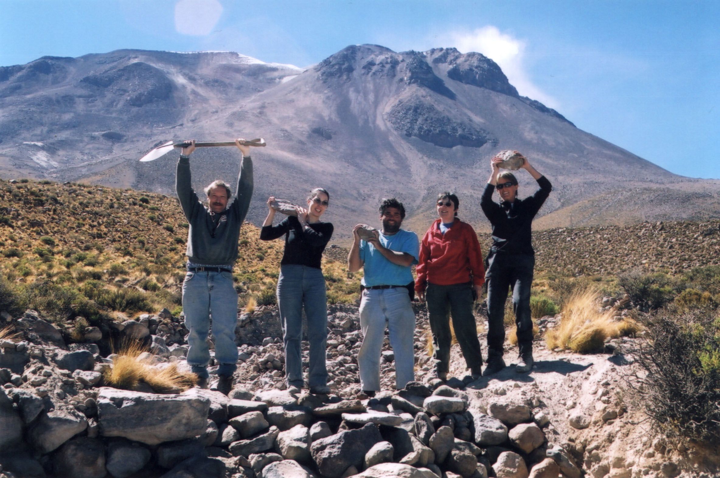 Stephen and four colleagues and students stand on a rocky road in front of a mountain, holding spades and rocks in the air above their heads