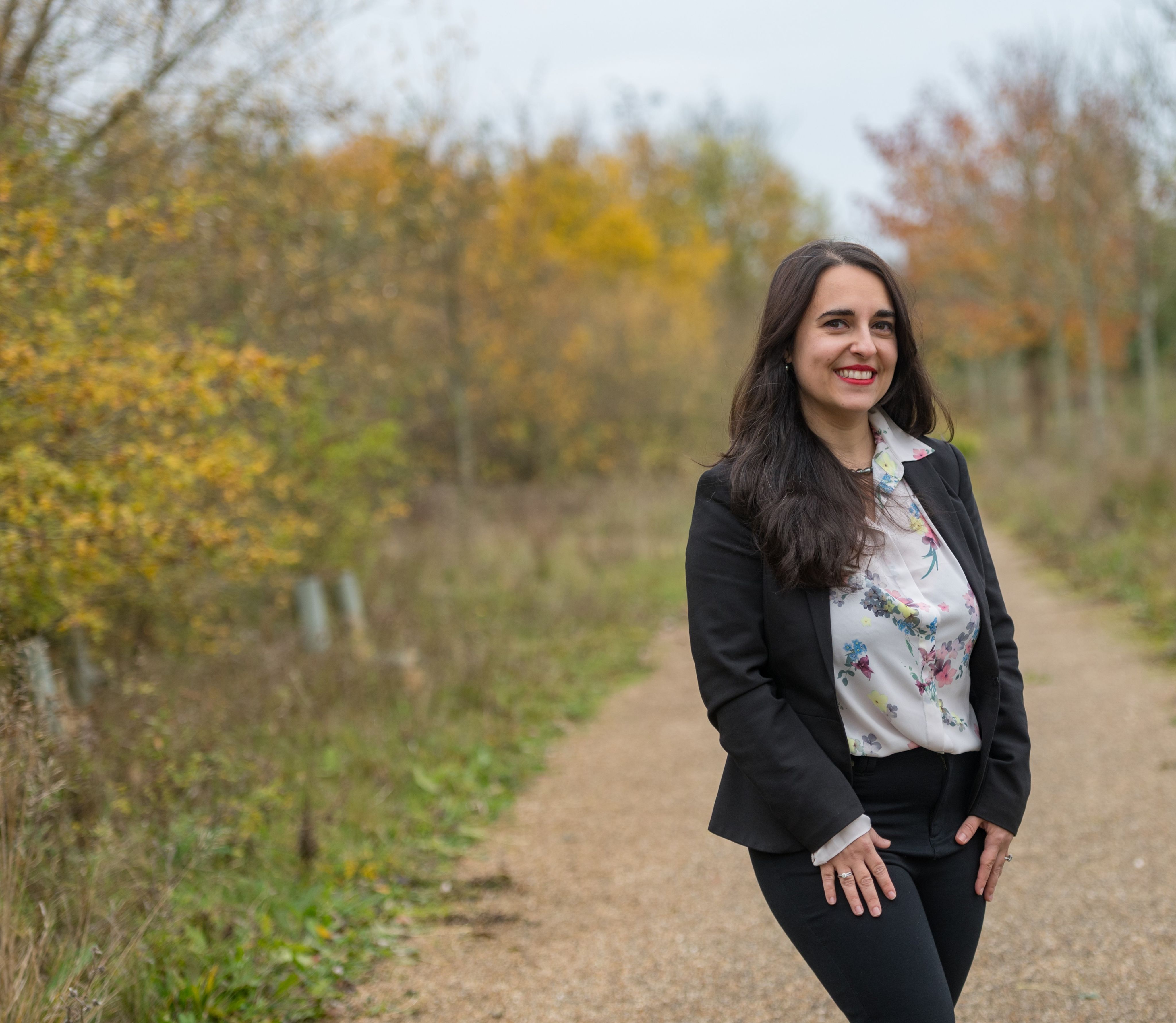 Giuliana standing in a garden outdoors, the leaves on the trees are blurred but are an autumnal colour. She wears a black blazer over a floral shirt, her hair is down