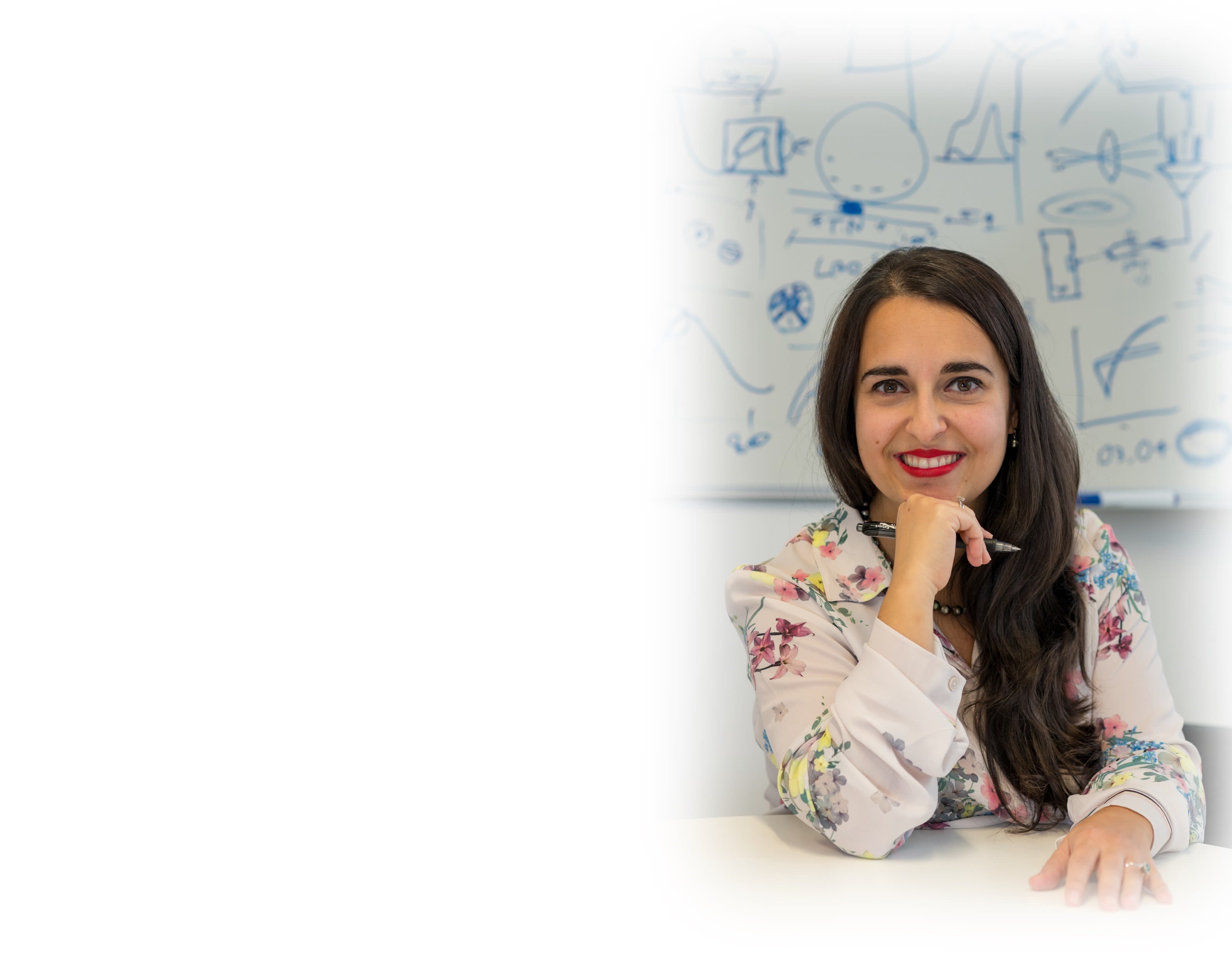 Giuliana sitting in front of a whiteboard filled with working and symbols. She is holding a pen with the same hand that is touching her chin as she poses for the picture smiling.