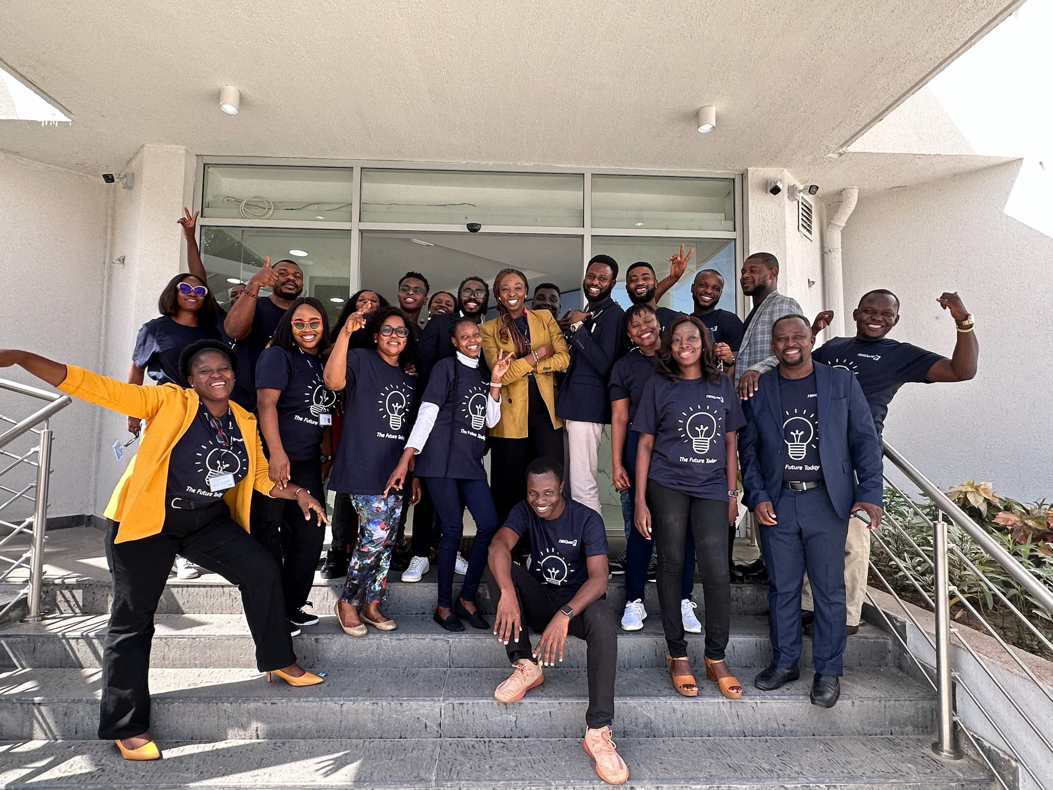 Lola with a group of over a dozen colleagues as they cheer for a photo, they are all wearing the same navy t-shirts with a lightbulb icon on it and a slogan that reads: 'The Future Today'