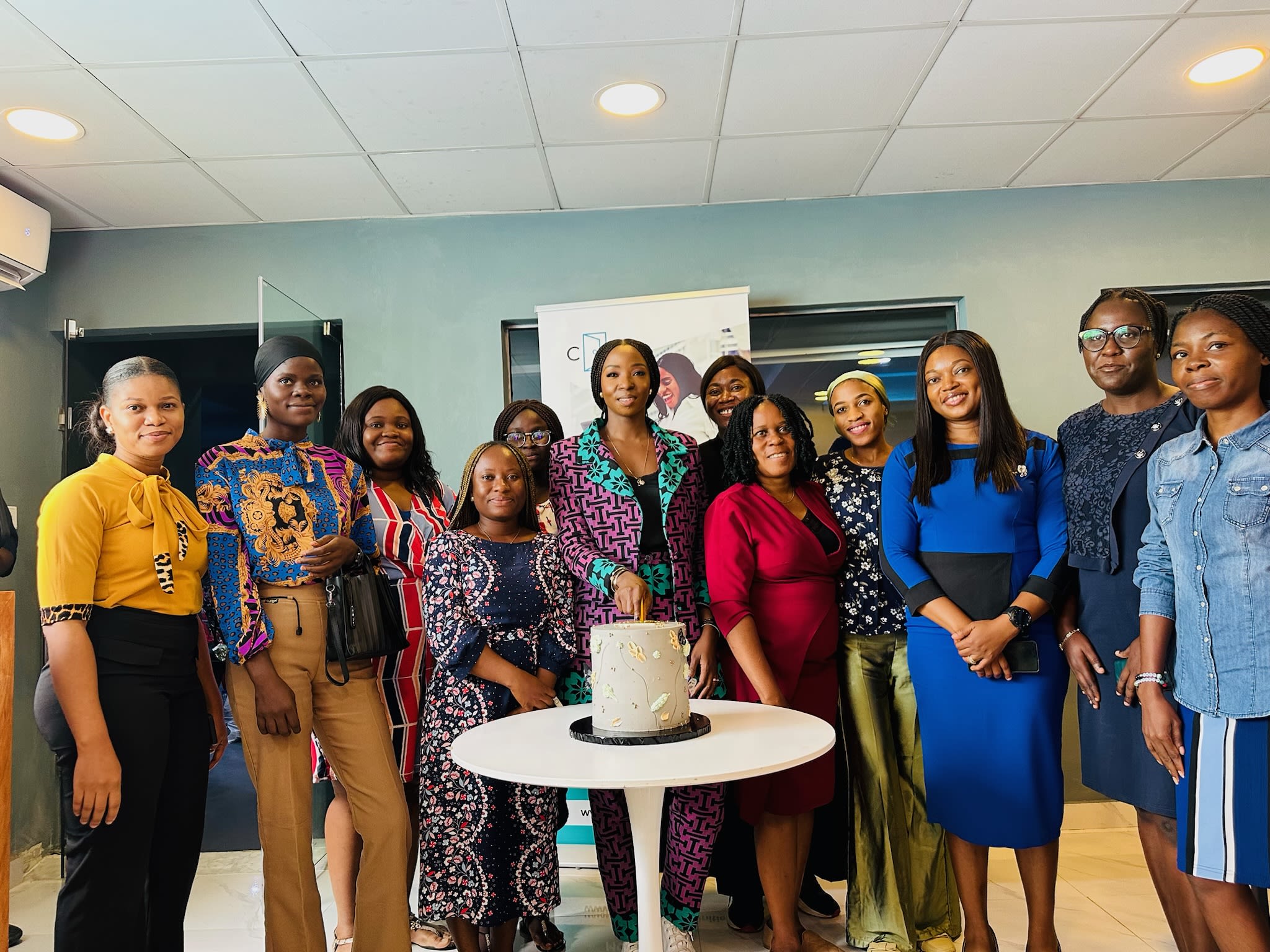 Lola pictured in the centre of a group of women. They all stand around a cake that has been placed on a small circular table. The setting looks corporate - like a classroom or meeting room 