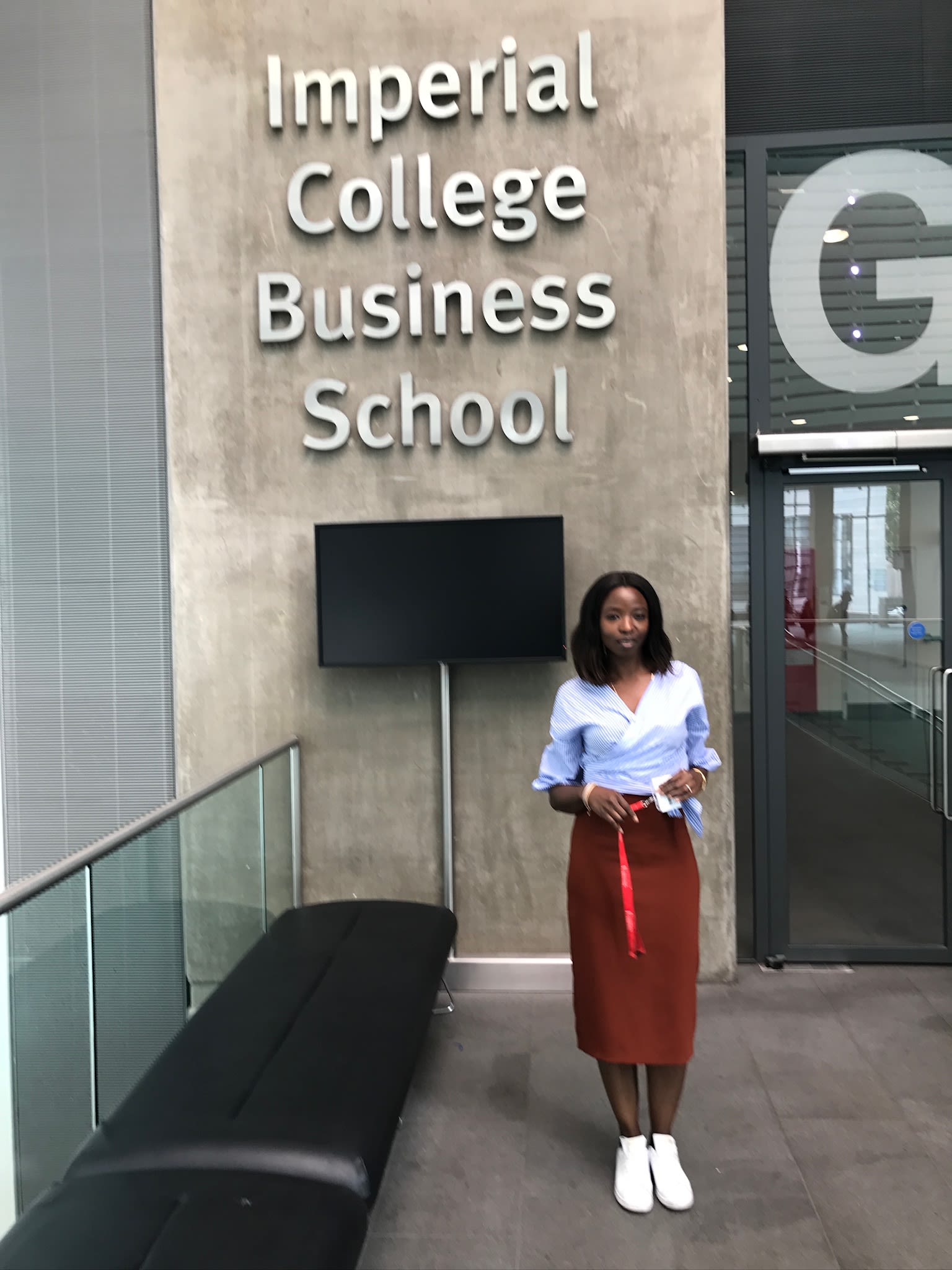 Lola is pictured standing outside of the large lettered sign that reads Imperial College Business School. She wears a smart outfit consisting of a shirt and a maxi skirt with white sneakers