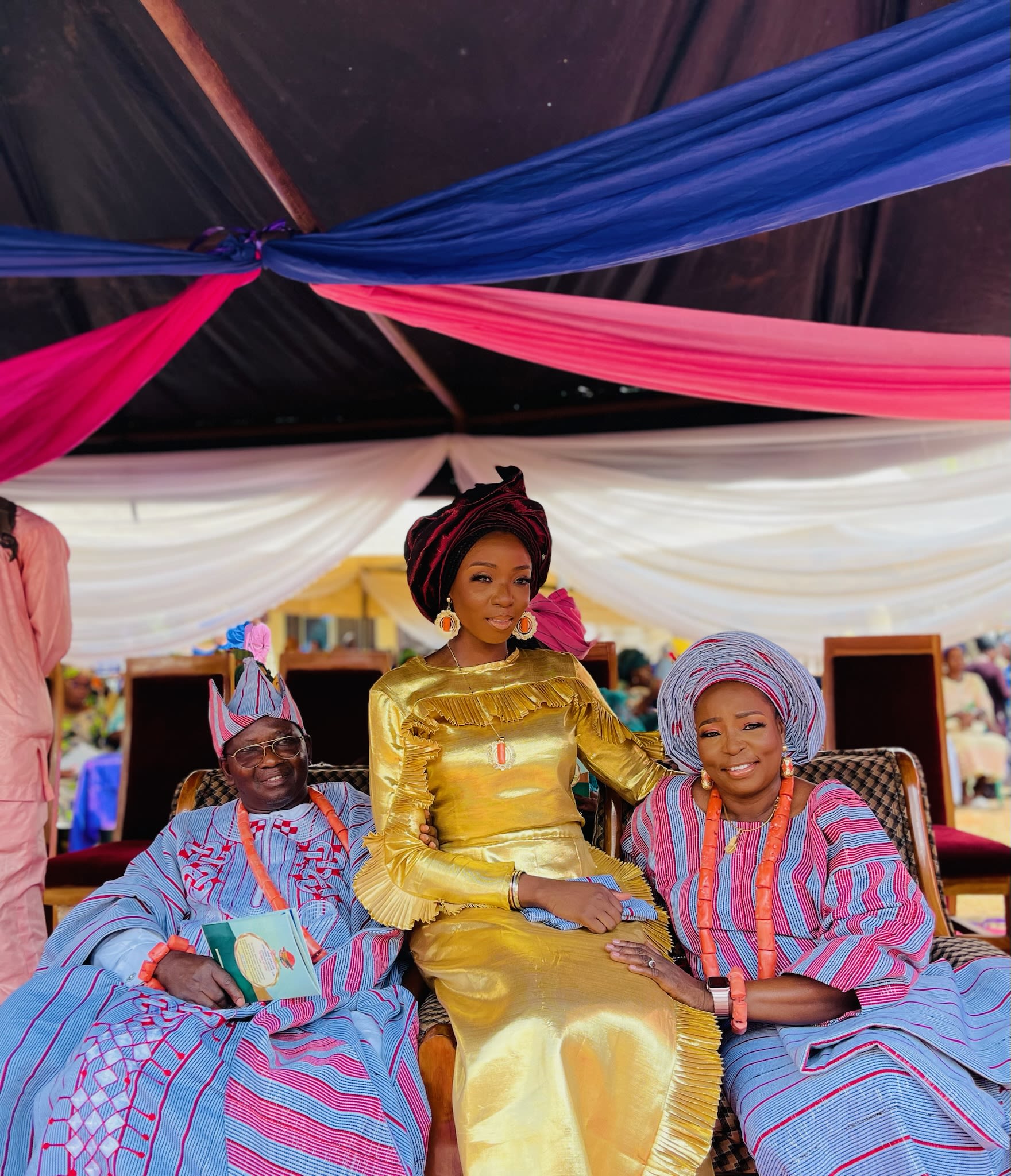 Lola sits in the middle of her two parents. They are wearing traditional ceremony wear. Lola wears a silky yellow long dress and her parents wear pink and blue traditional ensembles. They all wear headwear. Lola and her mum both wear chunky drop earrings to complement their outfits.