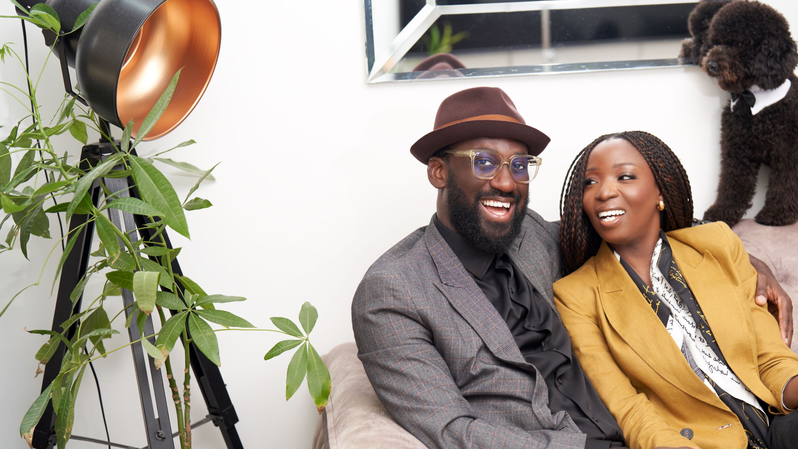 Lola with her partner, they are on a couch smiling at eachother. He has his arm around her. He wears glasses and a hat, whilst she wears a mustard yellow blazer and her long hair is braided.