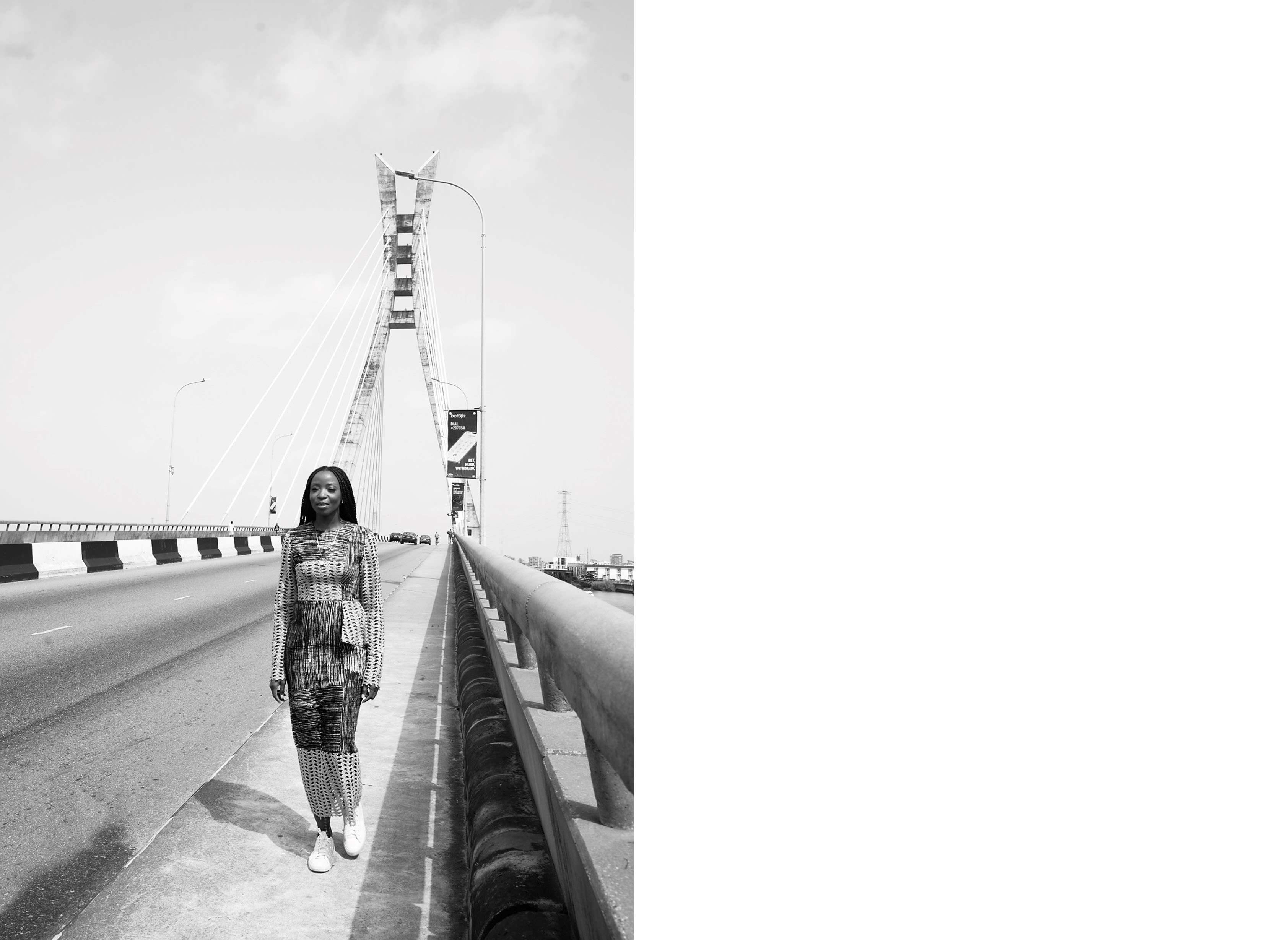 Lola walking purposefully towards the camera, she stands on a bridge on a bright day in Lagos. She is wearing a purple and black patterned maxi dress with white sneakers.