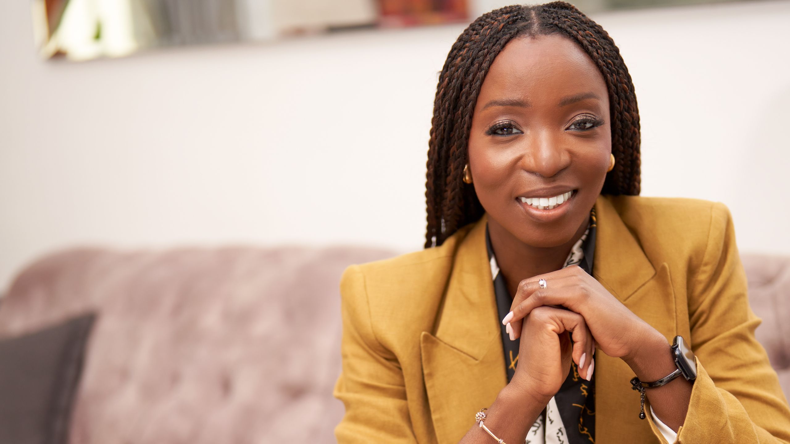 Lola with her hands clasped beneath her chin. She is smiling and wearing a mustard yellow blazer. She wears earrings, a watch and has a wedding ring visible.