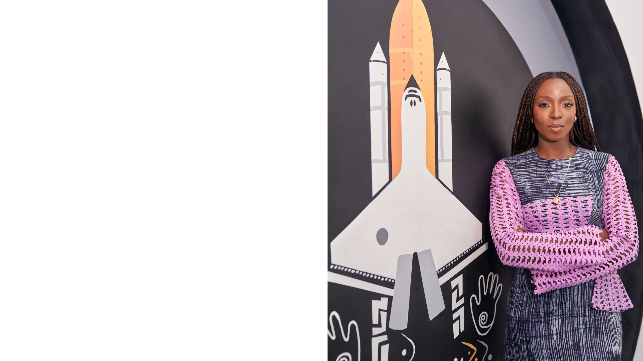 Lola stands against a mural of a rocket, she is wearing a colourful purple and black long sleeve top and stands with her arms crossed facing the camera