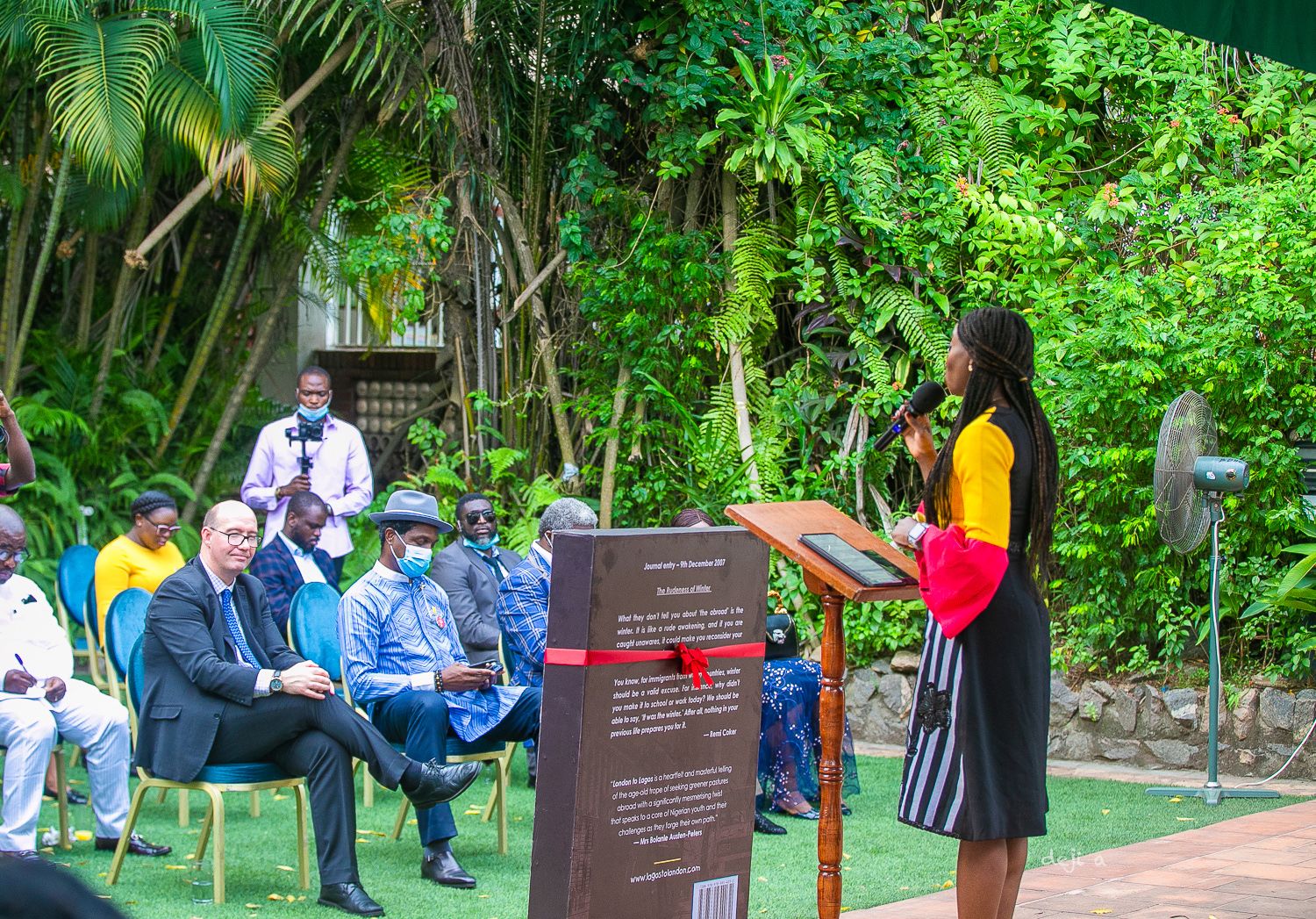 Lola speaks at a podium in a lush garden setting with what appears to be a select audience