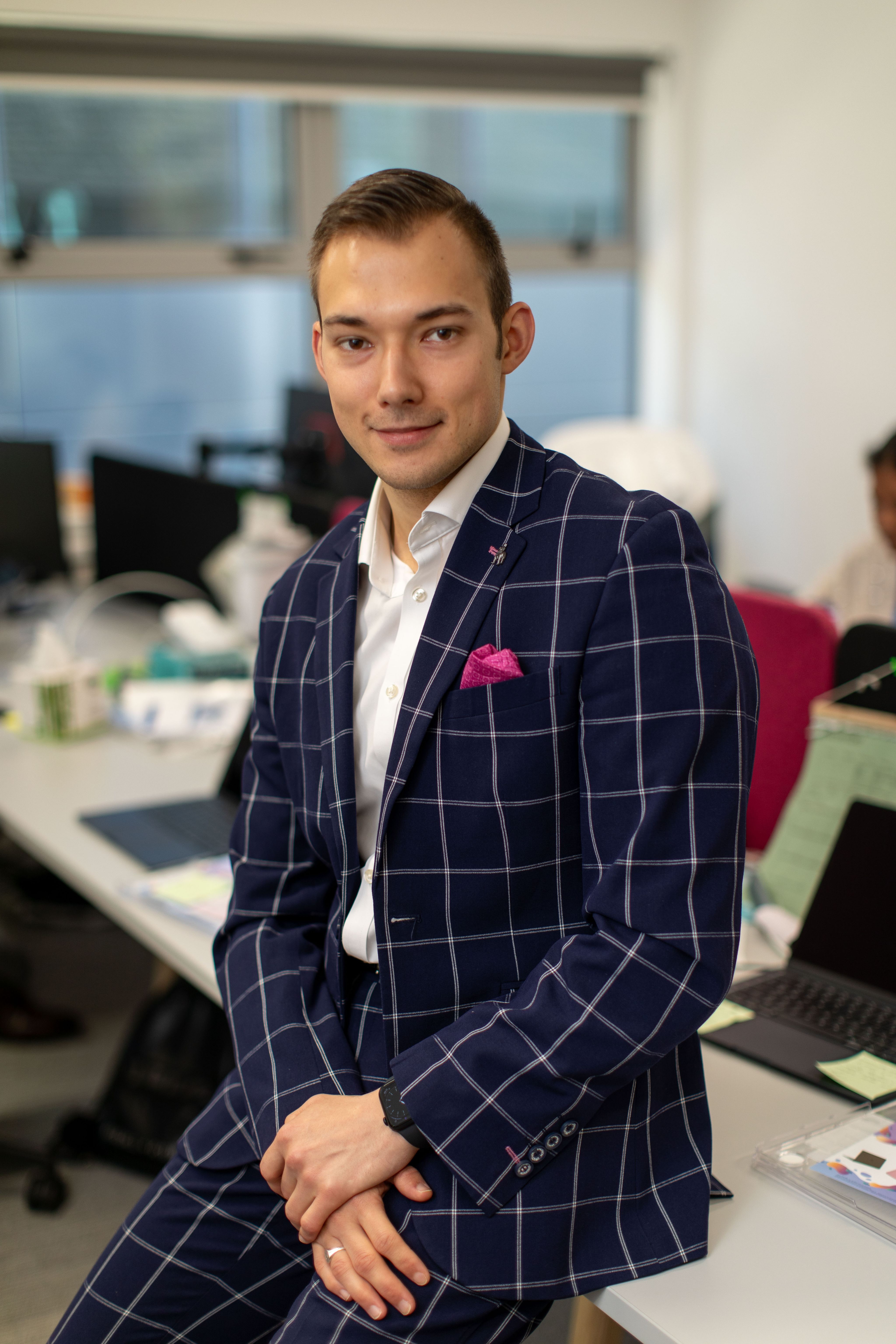 Miroslav wearing a navy checked suit, he leans casually against a desk and smirks at the camera