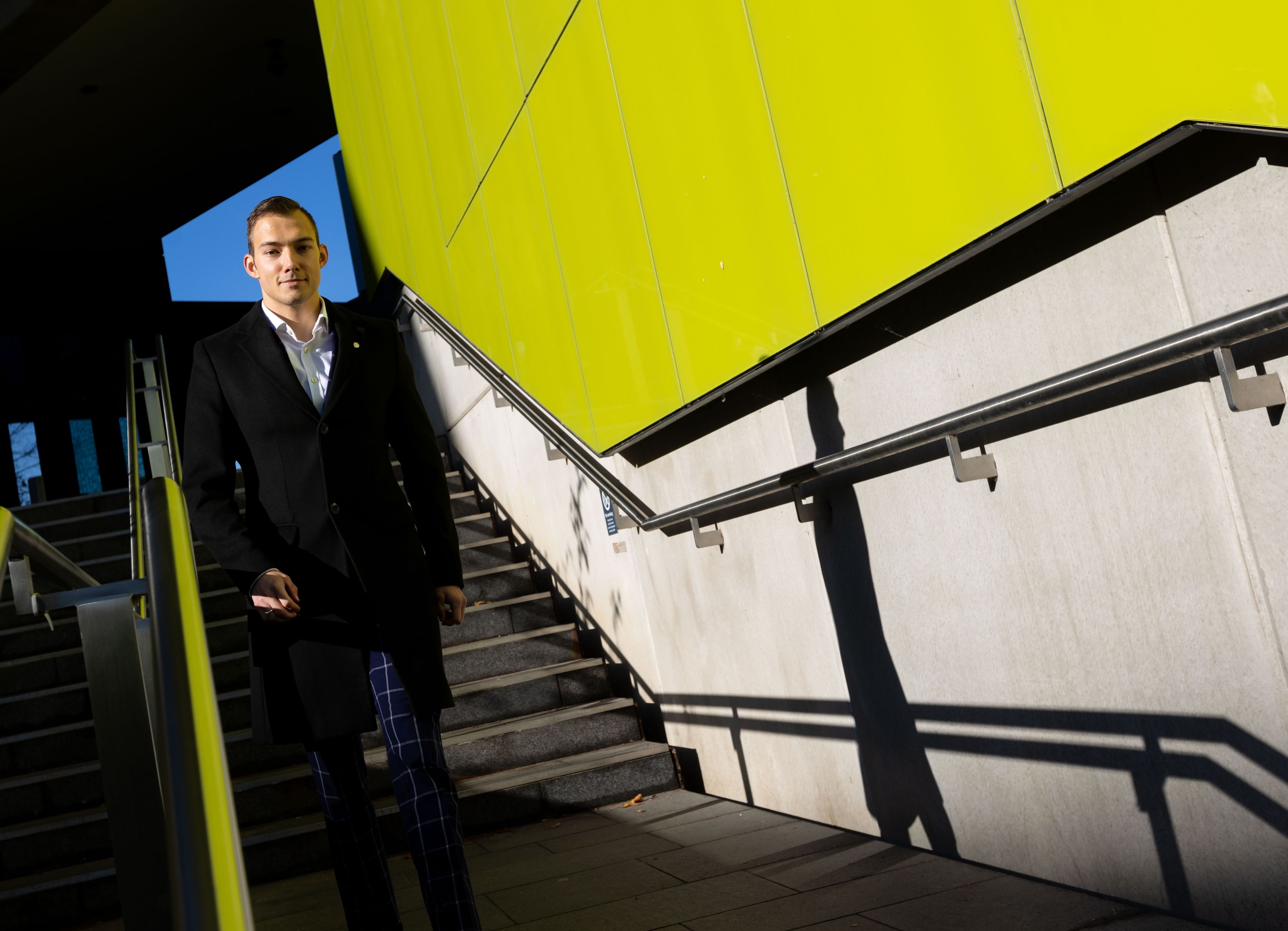 Miroslav walking down an illuminated stairwell. The walls are bright yellow. He wears a collared overcoat over his checked suit and his walking towards the camera