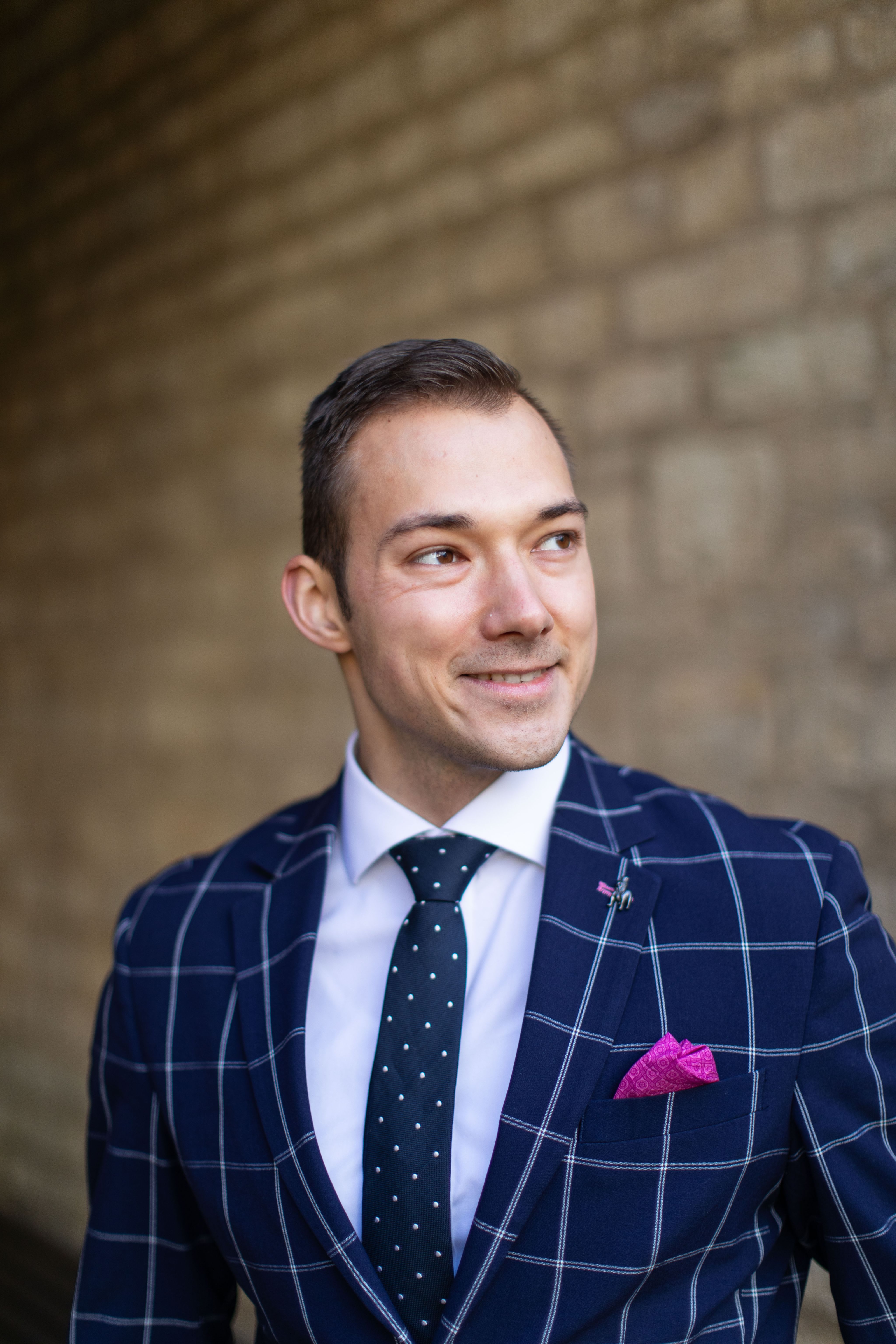 Portrait headshot of Miroslav, he is smiling and looking away from the camera, wearing a navy checked suit and standing against a brick wall that is photogenically blurred 