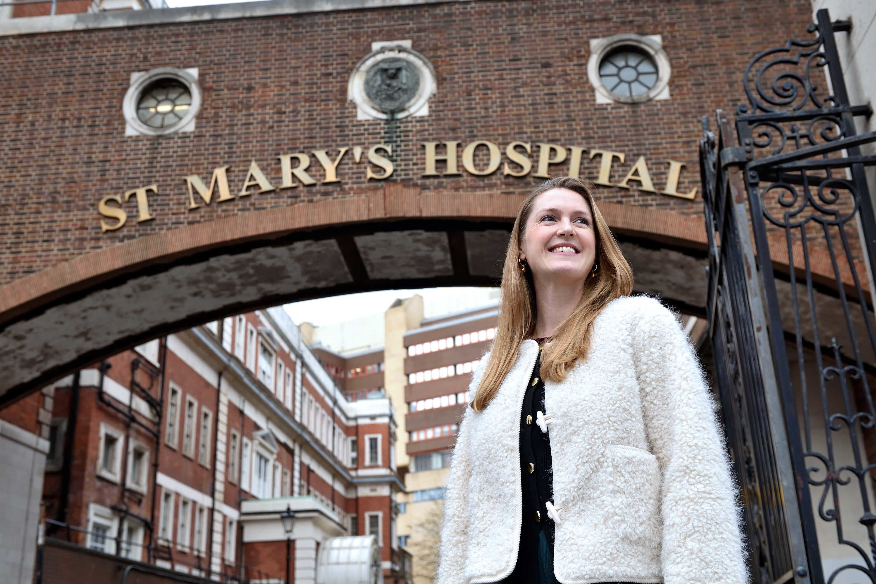 Anna wearing a white coat and smiling looking over the photographers right shoulder with the brown-brick St May's Hospital arch in the background